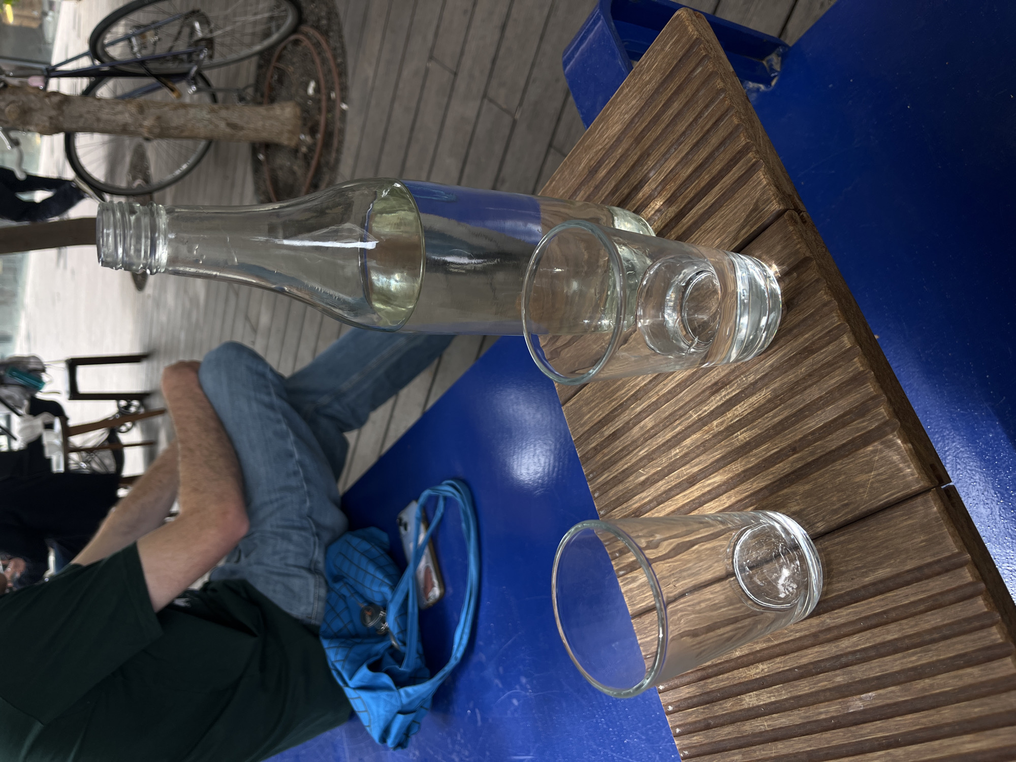 The image depicts a table setting at an outdoor cafe, likely in Tel Aviv, Israel. The table is composed of a wooden slat surface with a blue painted base. A partially empty glass bottle is lying horizontally on the table, alongside two empty drinking glasses. A person is seated at the table wearing jeans and a green shirt. A blue bag and a smartphone are placed on the blue table surface. In the background, a bicycle is leaning against a tree. The location is likely outdoors on a wooden deck.