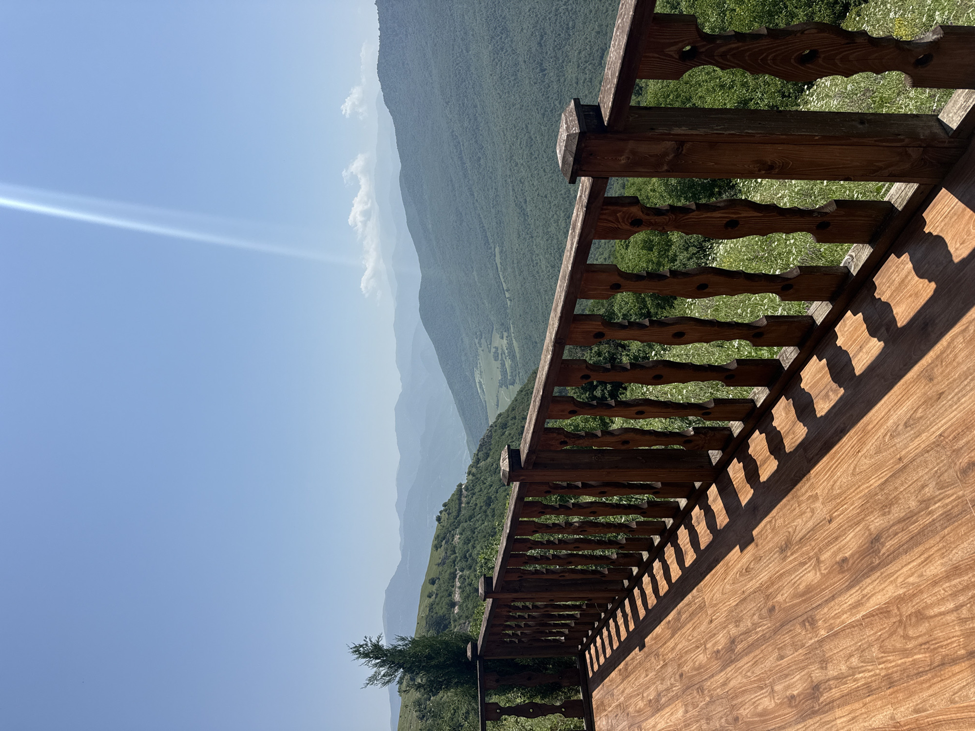 An outdoor perspective shot shows a wooden deck with a wooden railing on the left side. The railing is made of dark brown wood with ornate carvings on each spindle. The deck is made of polished wood planks with a light brown grain. Beyond the railing, a lush green valley stretches out, dotted with trees and wildflowers. In the distance, layers of mountains fade into a hazy blue, and the sky above is a clear, bright blue with a single, bright beam of sunlight shining from the upper right corner. The scene is likely captured from a vantage point in or near Yenokavan, Armenia, a location known for its natural beauty and mountainous landscape. There are no people or animals visible in the image.