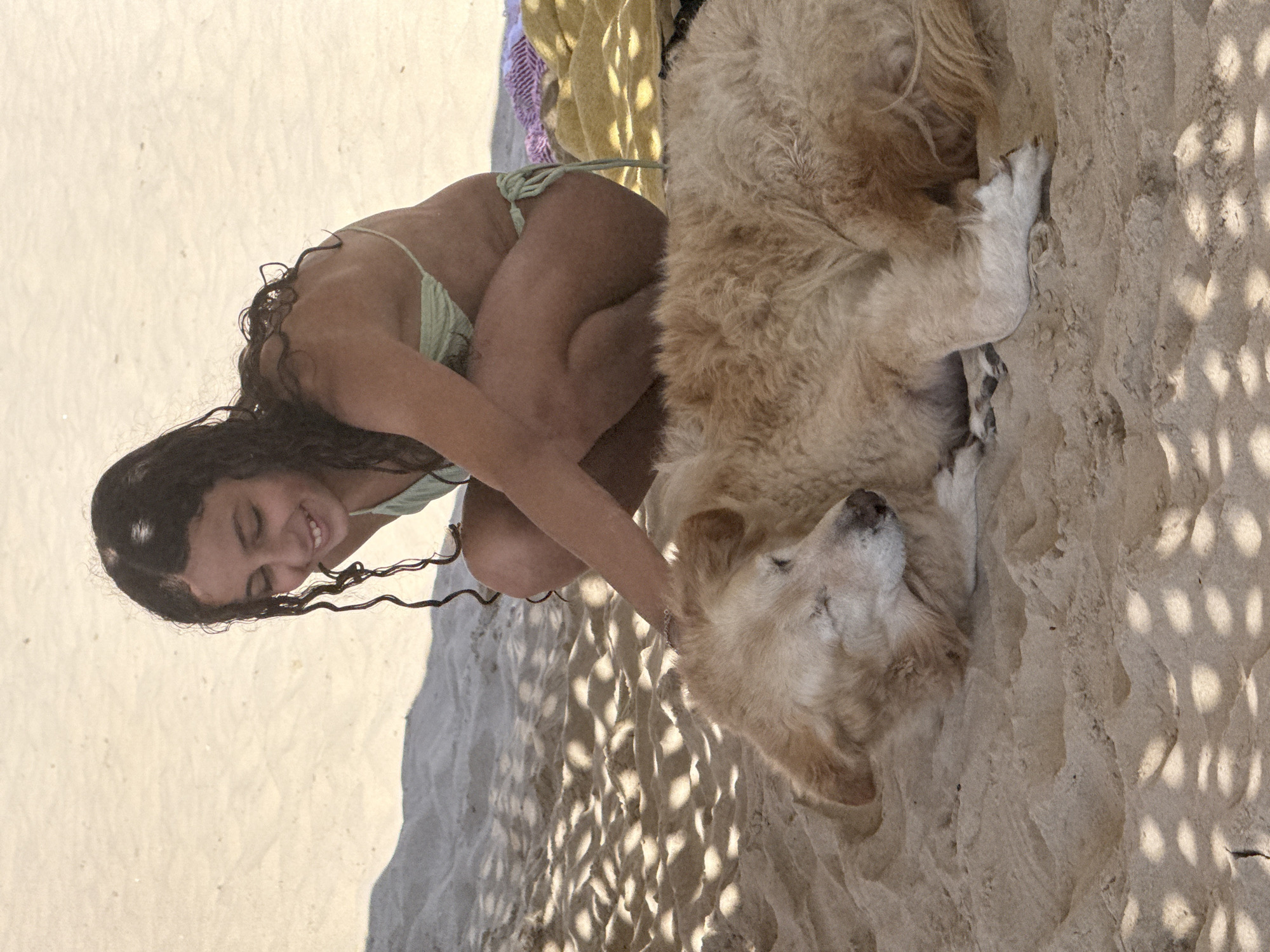 The image shows a woman and a dog on a sandy beach. The woman, with dark curly hair and wearing a light green bikini, is crouched down and appears to be interacting with the dog, a golden retriever or similar breed. The dog is lying down with its eyes closed, seemingly relaxed or sleeping. The background consists of light-colored sand, with dappled sunlight and shadow patterns from nearby foliage. The setting is likely a beach in Tel Aviv, Israel, as that information was provided. Other visible elements include a partially visible towel or blanket in the background.
