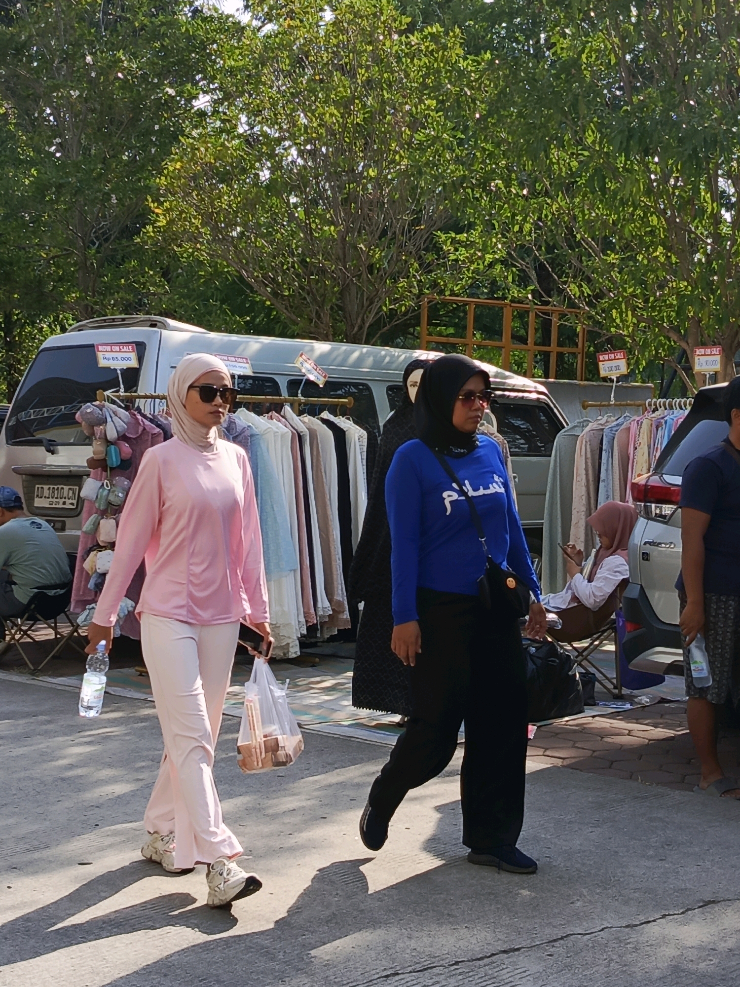 The image captures an outdoor scene, likely a street market, in Tangerang, Indonesia. Two women are prominently featured walking on a paved surface. The woman on the left wears a pink hijab and matching pink outfit, carrying a water bottle in her right hand and a plastic bag containing some items in her left. She is also wearing sunglasses and white sneakers. The woman on the right is dressed in a black hijab and a blue long-sleeved shirt with Arabic writing on it, paired with black pants and dark shoes. She has a black crossbody bag.
Behind them, a white van is parked, with clothing racks displaying various garments. Signs indicating "NOW ON SALE" with prices are visible on the van. Further in the background, trees provide shade, and other people can be seen, including a person sitting in a chair next to a rack of accessories, and another person partially visible to the right. The overall impression is of a bustling marketplace under natural daylight.