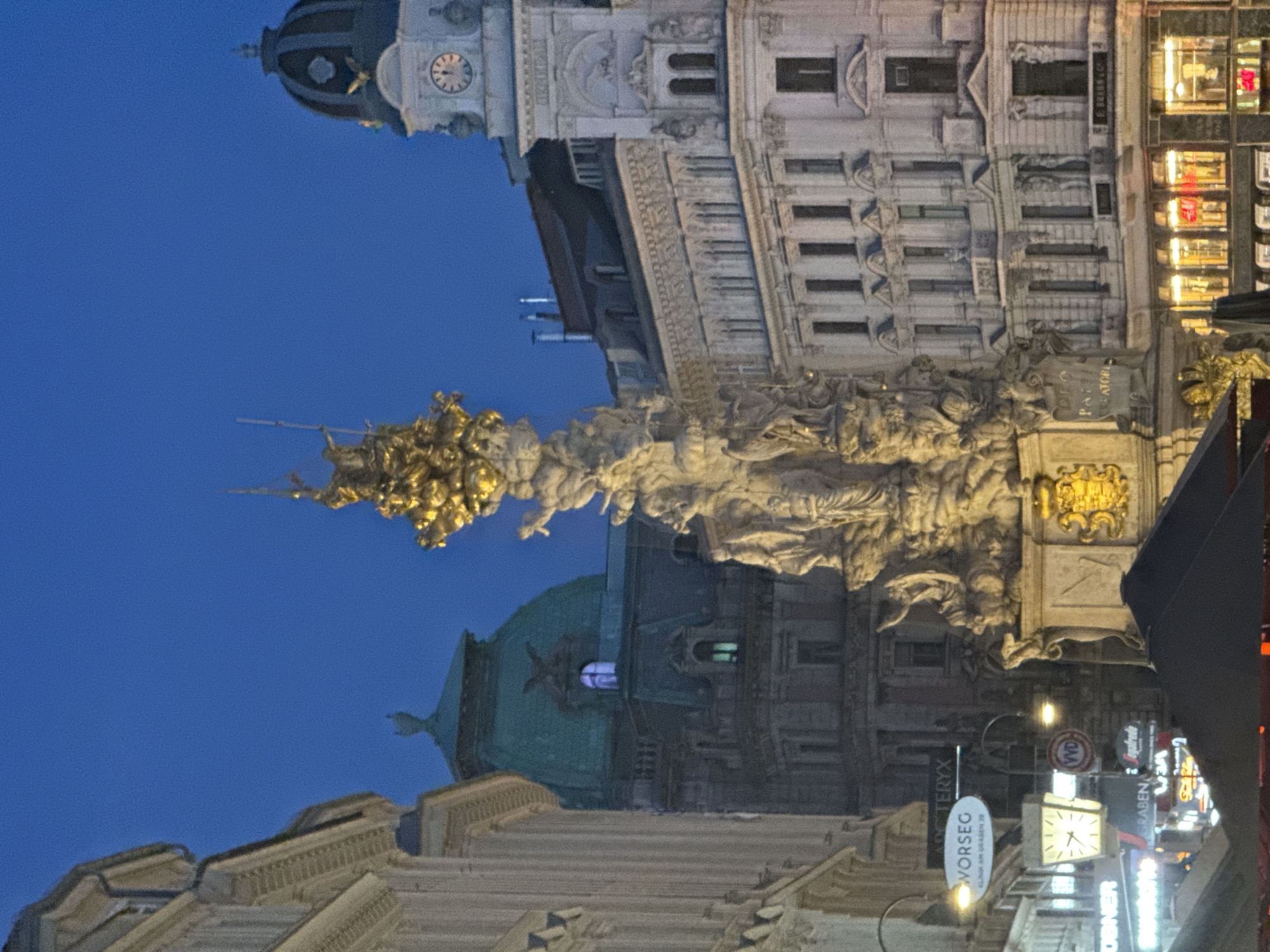 This is a low-angle, nighttime photograph of an elaborate architectural monument, likely a fountain or memorial, in Vienna, Austria. The central subject is a complex sculpture depicting multiple figures and mythical creatures in ornate gold and stone. Above and behind the sculpture, several imposing buildings with detailed facades and illuminated windows are visible, suggesting an urban setting. One building features a clock tower with a visible clock face, indicating it's likely evening. The sky is a deep, clear blue, typical of a twilight or night sky.
The monument itself appears to be the focal point, with the figures carved in dynamic poses. The overall scene evokes a sense of grandeur and historical significance. To the right of the monument, there is a modern building with glowing windows, contrasting with the older architecture. Several signs are visible, including "WORSEG", "TER", and other text that is partially obscured or difficult to read due to the lighting and angle. The overall impression is one of historical architecture illuminated against a dark sky, with the sculpture serving as a dramatic centerpiece.