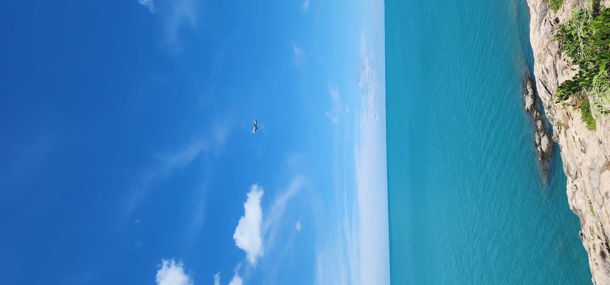 This media file captures a serene, tropical scene. The primary subjects are the vast, vibrant turquoise ocean meeting a rocky, vegetated coastline under a clear blue sky with scattered white clouds. An airplane is visible in the sky, appearing small and distant, suggesting it is either taking off or landing.
The scene depicts a tranquil day in what appears to be a coastal location. The clear blue sky indicates fair weather, likely during daylight hours, given the brightness of the scene. The presence of lush green vegetation on the rocky cliff face suggests a warm climate. The calm water of the ocean implies either a peaceful day or a sheltered bay. The airplane introduces an element of human activity within the natural landscape.
Notable details include the stark contrast between the deep blue of the sky and the bright turquoise of the sea. The texture of the rocky cliff is also apparent, with its rugged surface and scattered greenery. The smallness of the airplane against the expansive sky and sea emphasizes the grandeur of nature. No text is visible in the media file.
Considering the provided location context of Baan Chaweng Noi, Thailand, this scene likely represents a typical view of the coastline, potentially from a high vantage point on the cliff. The calm, clear waters and sunny skies are characteristic of this region. The airplane could be a passenger jet arriving at or departing from the nearby Samui Airport. The overall mood is one of peace and natural beauty.