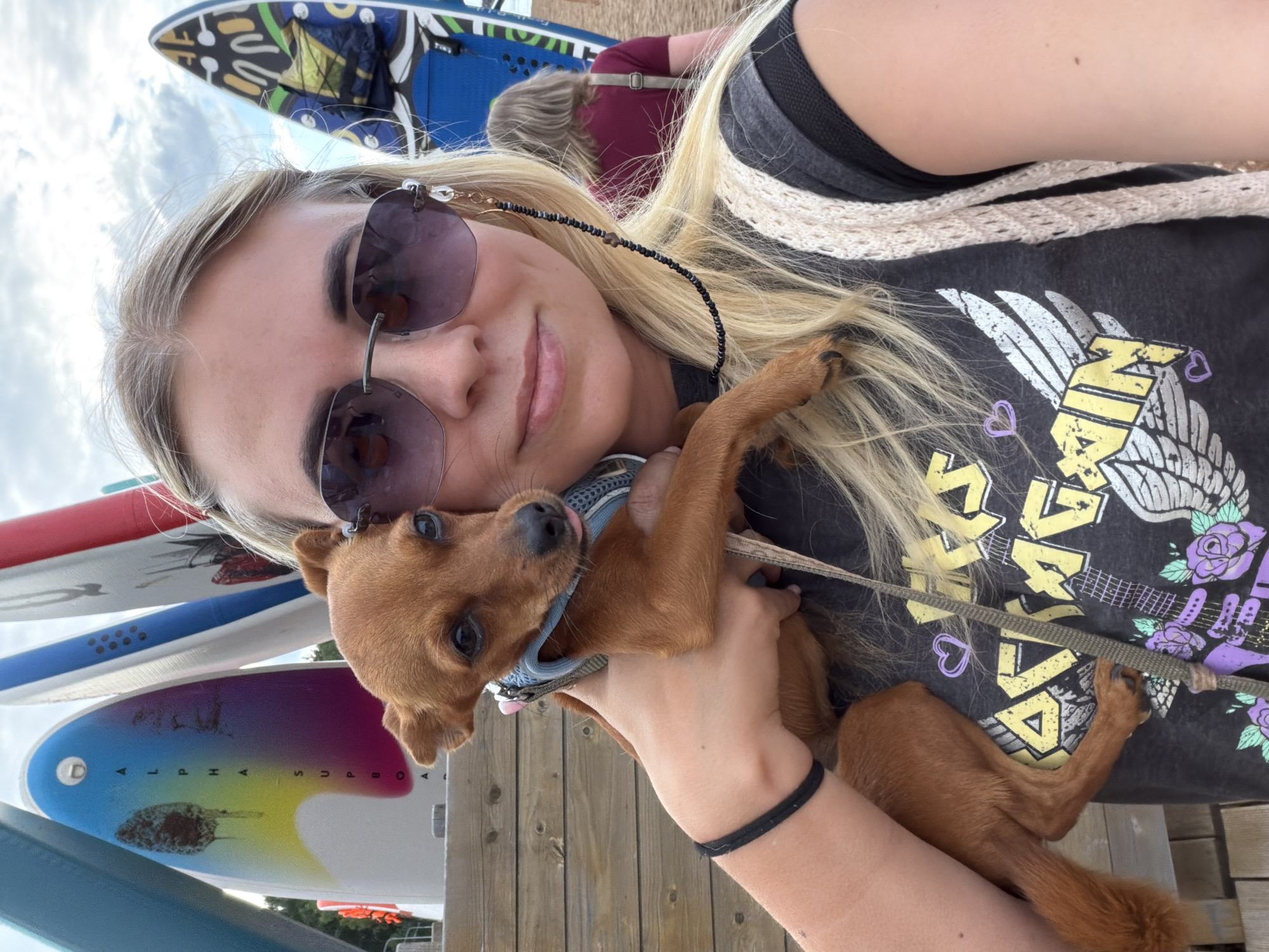 A young woman with blonde hair is taking a selfie with her small, brown dog. She is wearing sunglasses and a black t-shirt with a graphic design featuring wings, a guitar, and flowers, as well as some text. The dog is wearing a blue harness and a leash. The woman is holding the dog close, and both appear happy. In the background, there are several colorful surfboards or paddleboards stacked vertically. The setting appears to be outdoors, possibly near a beach or a waterfront, with a wooden deck visible beneath them. The lighting suggests it is daytime, with a bright sky.