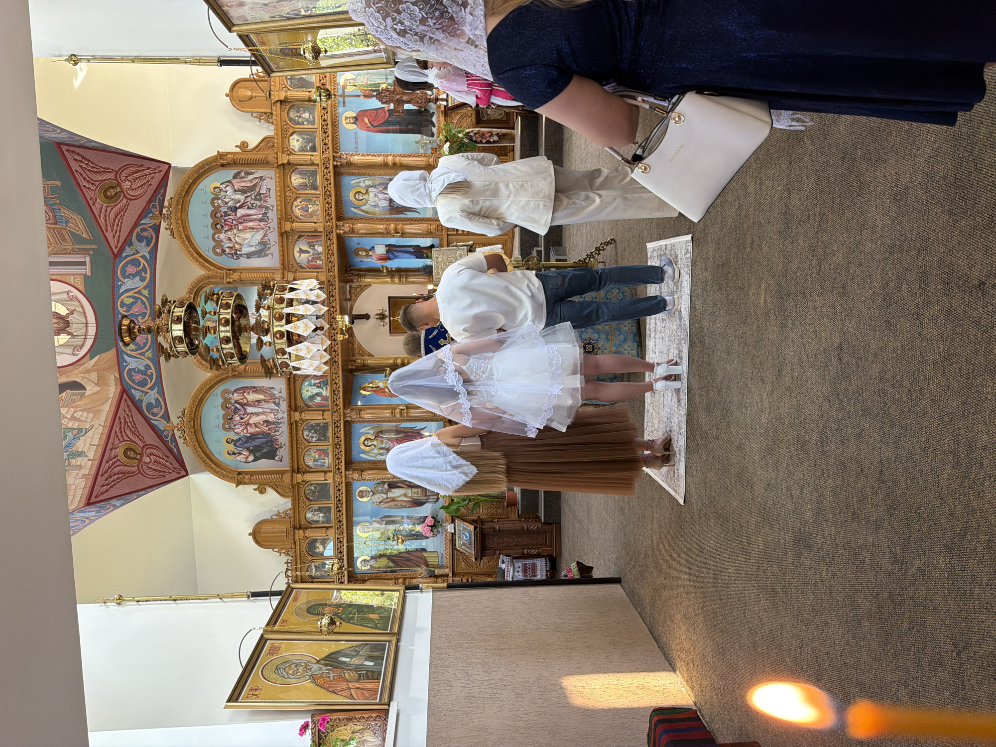 This image depicts an indoor scene, likely within an Orthodox church in the city of Strășeni, Moldova. In the foreground, the right side of a woman wearing a dark blue dress and a white lace head covering is visible, holding a white handbag. To her left, a man in a white t-shirt and jeans stands facing an altar. Beside him, a woman in a white wedding dress and veil, and another woman in a brown dress and white veil, also face the altar. The altar area itself is ornately decorated with icons and religious imagery. A large, multi-tiered chandelier hangs from the ceiling, and the walls and ceiling are adorned with frescoes. A candle in the lower left corner of the image is lit, casting a warm glow. The floor is carpeted. The overall atmosphere suggests a religious ceremony, possibly a wedding.
