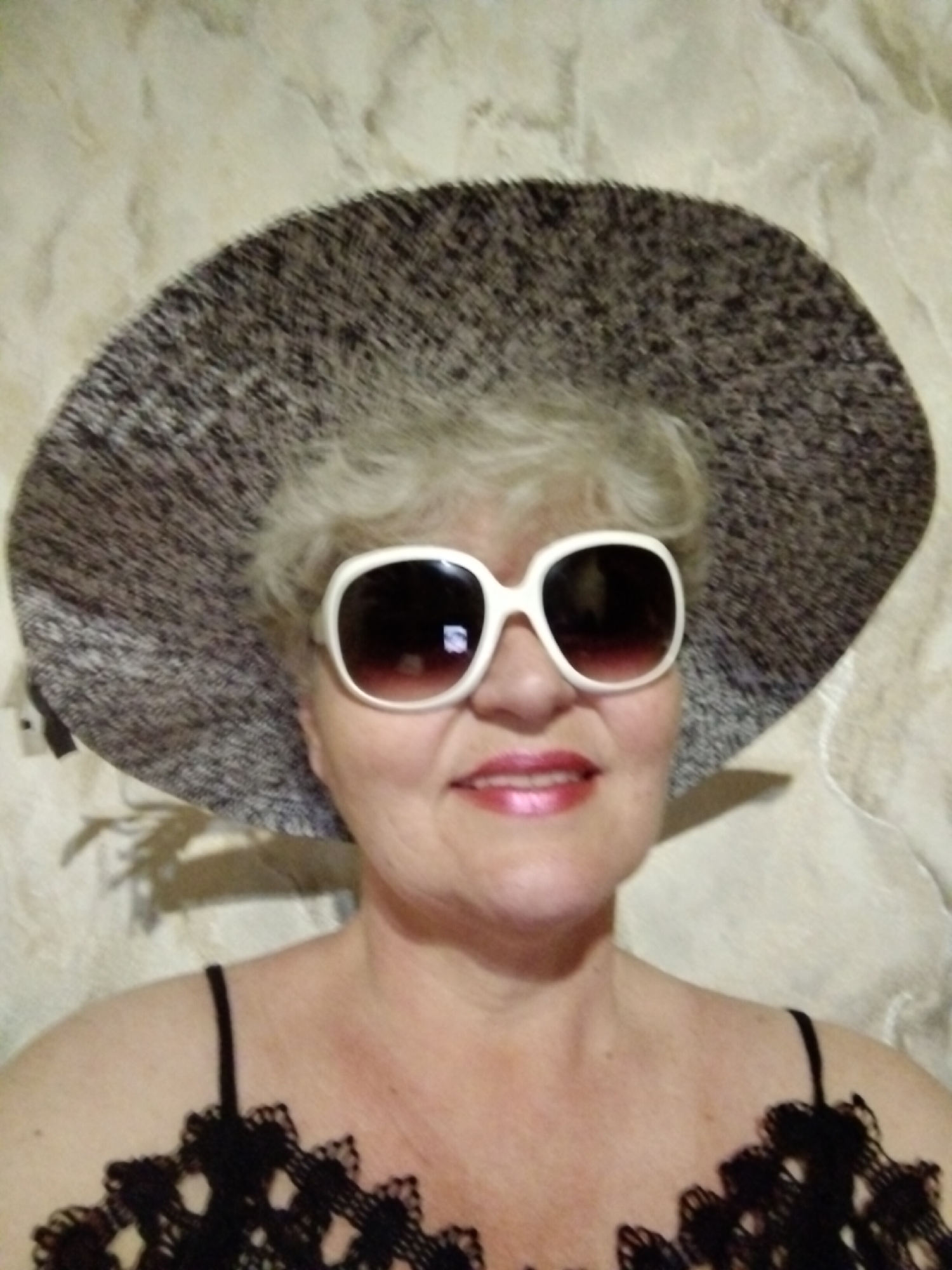 A woman with short, gray hair is wearing a large, speckled hat and oversized white sunglasses. She has lipstick on and is wearing a black top with lace detailing. The background appears to be a neutral, patterned wall. There is no information to conclude this image was taken in Horishni Plavni, Ukraine.