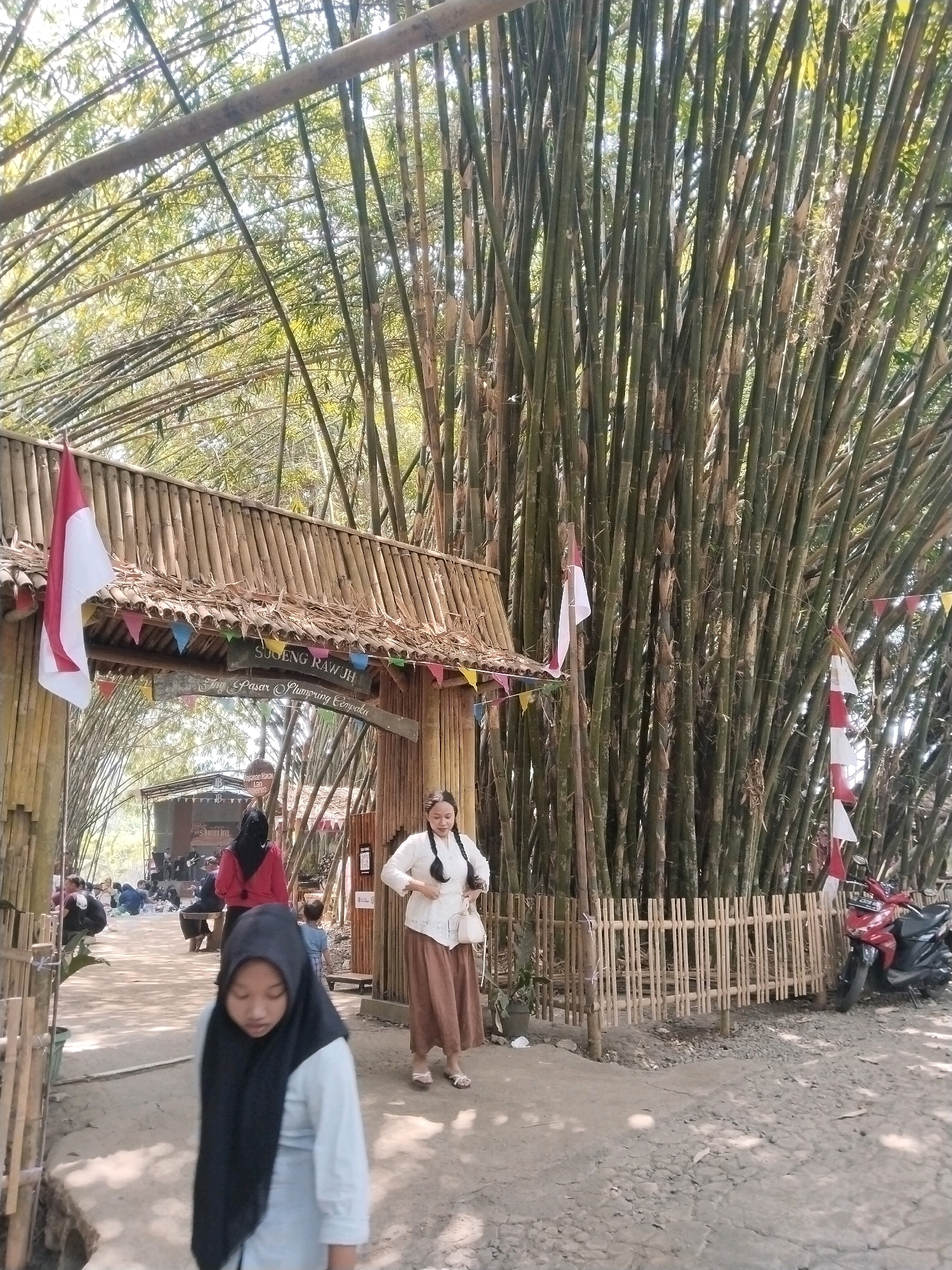 The image depicts an outdoor scene at a bamboo forest park, likely in Cempaka, Indonesia, identified by the sign above the entrance which reads "SUGENG RAWUH". The entrance itself is a rustic structure made of bamboo with a thatched roof, adorned with Indonesian flags and colorful triangular pennants. Tall, dense bamboo stalks dominate the background, creating a natural canopy with sunlight filtering through.
In the foreground, a young girl wearing a black hijab and a light blue shirt is out of focus. To her right, a woman with her hair in braids, wearing a white traditional top and a long brown skirt, walks past. She carries a white purse. Further back, a stage area is visible with several people seated, some appearing to be performers. Other visitors are scattered around, with some seated on the ground. A red motorcycle is parked near a bamboo fence to the right of the frame. The ground is a mixture of dirt and gravel. The overall atmosphere suggests a local gathering or event within a natural setting.