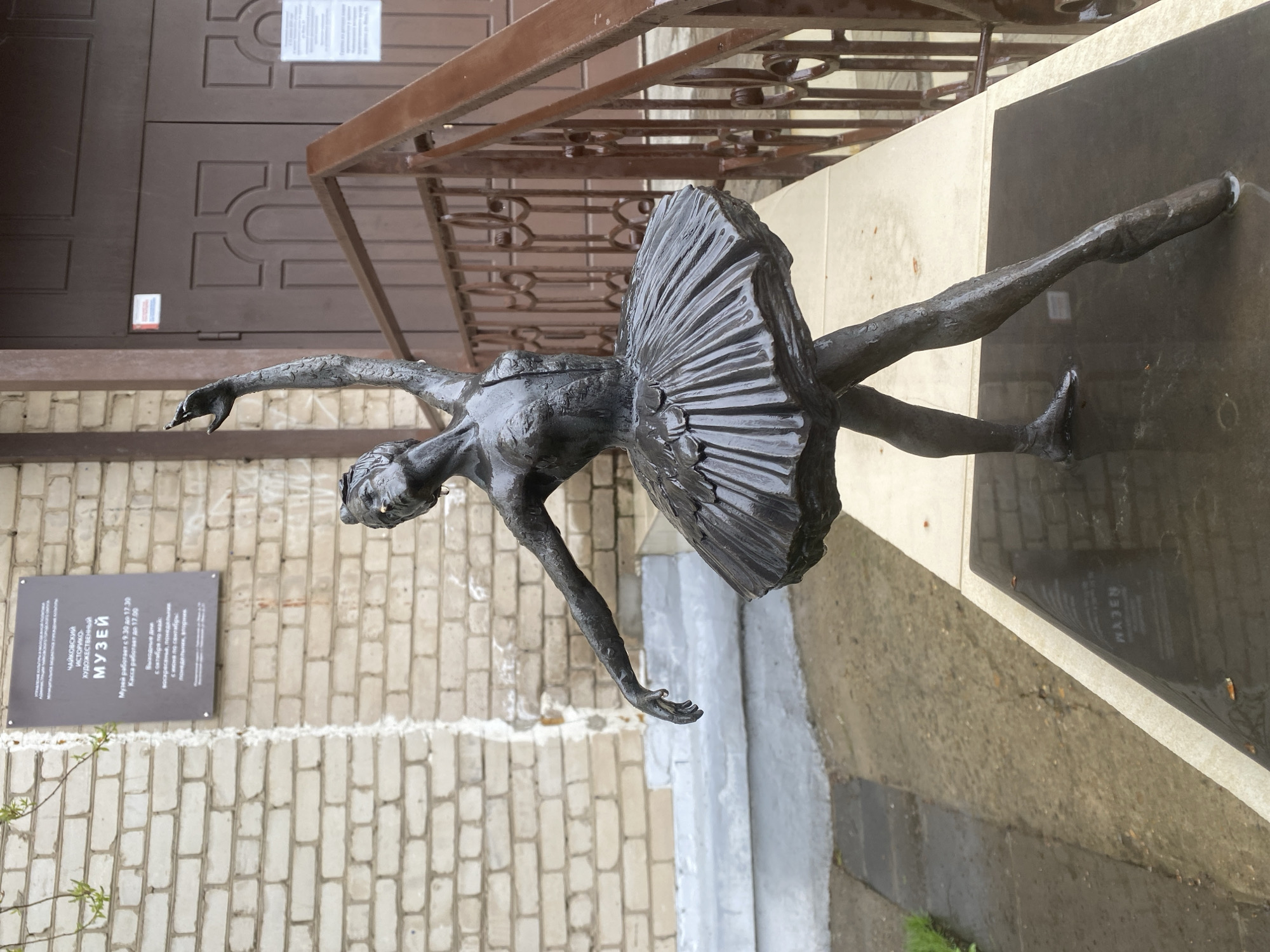An outdoor, full-length bronze statue of a ballerina is positioned vertically in front of a building facade in Chaikovsky, Russia. The statue depicts a ballerina in a graceful, extended pose, with one leg lifted high behind her, arms outstretched, and head tilted upwards. Her tutu is sculpted with fan-like pleats, and the entire sculpture appears wet, with visible droplets on its surface, suggesting recent rain or damp weather.
The statue rests on a dark, reflective base, which also appears wet. The base is situated on a light-colored, possibly concrete, ledge. Behind the statue, a light-colored brick wall rises, with a dark brown, ornate metal railing running along the top of the ledge, separating the statue from the building entrance. A dark brown door with decorative geometric patterns is visible on the upper left, set into the brick wall. The overall lighting is diffused and even, indicative of an overcast day.
A rectangular sign is affixed to the brick wall to the left of the statue. The visible Russian text on the sign, when translated, reads:
"CHAIKOVSKY HISTORICAL AND ART MUSEUM"
"The museum is open from 9:30 to 17:30"
"The ticket office is open until 17:00"
"Days off:"
"from October to May: Sunday, Monday"
"from June to September: Monday, Tuesday"
A distorted reflection of the word "МУЗЕЙ" (MUSEUM), appearing as "WAJEN" in the OCR, is visible on the wet, dark base of the statue. The ground in front of the statue and base is concrete, also appearing wet. Small green plant shoots are visible near the lower left corner.