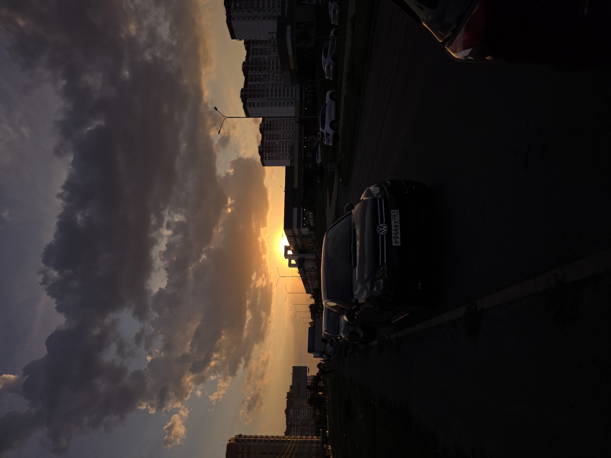 A vertical, low-angle shot captures a street scene in Rostov-on-Don, Russia during sunset. The sky is filled with dramatic, dark clouds interspersed with patches of bright orange and yellow, indicating the sun is low on the horizon, likely setting behind a building. Silhouetted against the vibrant sky are several multi-story residential buildings and streetlights. The foreground is dominated by the dark silhouettes of parked cars lining the street, with the license plate of a black Volkswagen SUV clearly visible as "P944BX 761 RUS". A few people are visible walking on the sidewalk in the distance. The overall atmosphere is moody and dramatic, characterized by the contrast between the dark foreground and the illuminated sky.