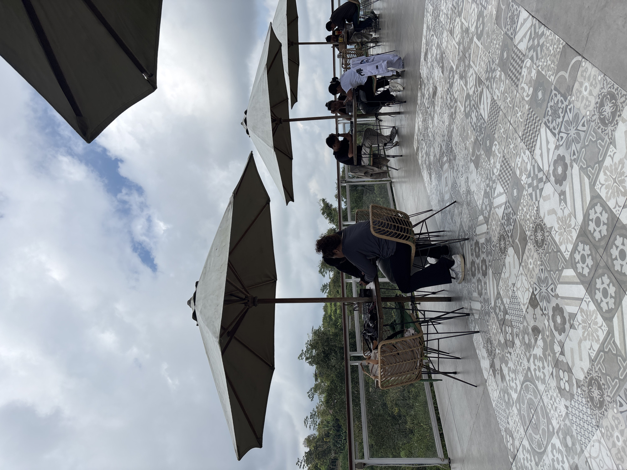 The image depicts an outdoor seating area in Baturiti, Indonesia. Several people are seated at tables under large, light-colored umbrellas. The floor is covered with decorative, patterned tiles. The area overlooks a lush, green hillside with trees. The sky is cloudy. Individuals are engaged in various activities, primarily sitting and interacting at the tables. Furniture consists of chairs with wicker elements. A glass barrier is visible, likely serving as a railing or windbreak. The atmosphere seems relaxed and casual.