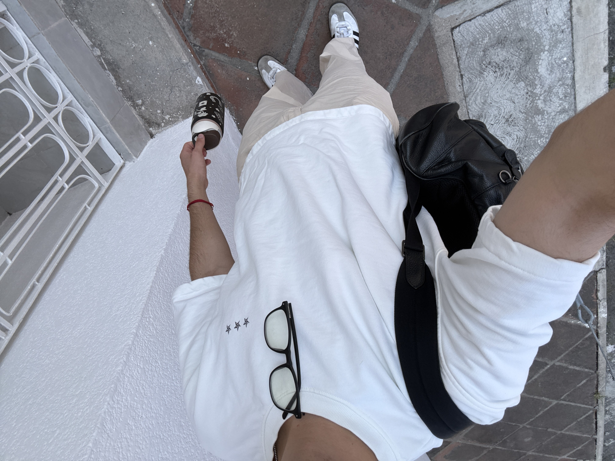 The image shows a person taking a selfie in what appears to be a street or sidewalk in Puebla, Mexico. The person is standing on a surface paved with patterned tiles and is holding a cup with a dark, patterned exterior and a brown base. They are wearing light-colored pants, white sneakers with gray accents, and a white T-shirt adorned with three star-shaped symbols. A pair of glasses is placed on the shirt, and a black leather backpack with a black strap is slung over the person's shoulder. A red string bracelet is visible on their wrist. The environment includes a white wall with a window featuring decorative ironwork. The lighting is soft and suggests daytime. The perspective is from a high angle, looking down at the person. The location is Puebla, Mexico.