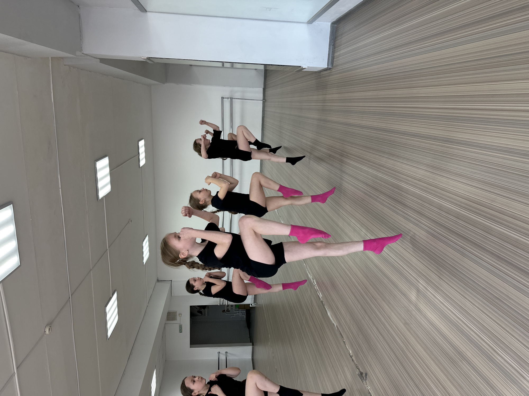 The image shows five young girls in what appears to be a dance or exercise studio. The location is likely in Votkinsk, Russia, due to the image being found online from that region. The room has a white ceiling with rectangular panel lights, white walls, and a floor with horizontal light brown and grey stripes. The girls are lying on the floor in various poses, all wearing black shorts and tops. Three of them have bright pink socks, while the other two have black socks. They all have their arms raised in front of them and their knees are in the air. There's a closed door with an inscription in Russian in the background on the left side of the frame. A horizontal pole is fixed to the wall behind the door, likely for ballet practices. The scene suggests a rehearsal or training session.