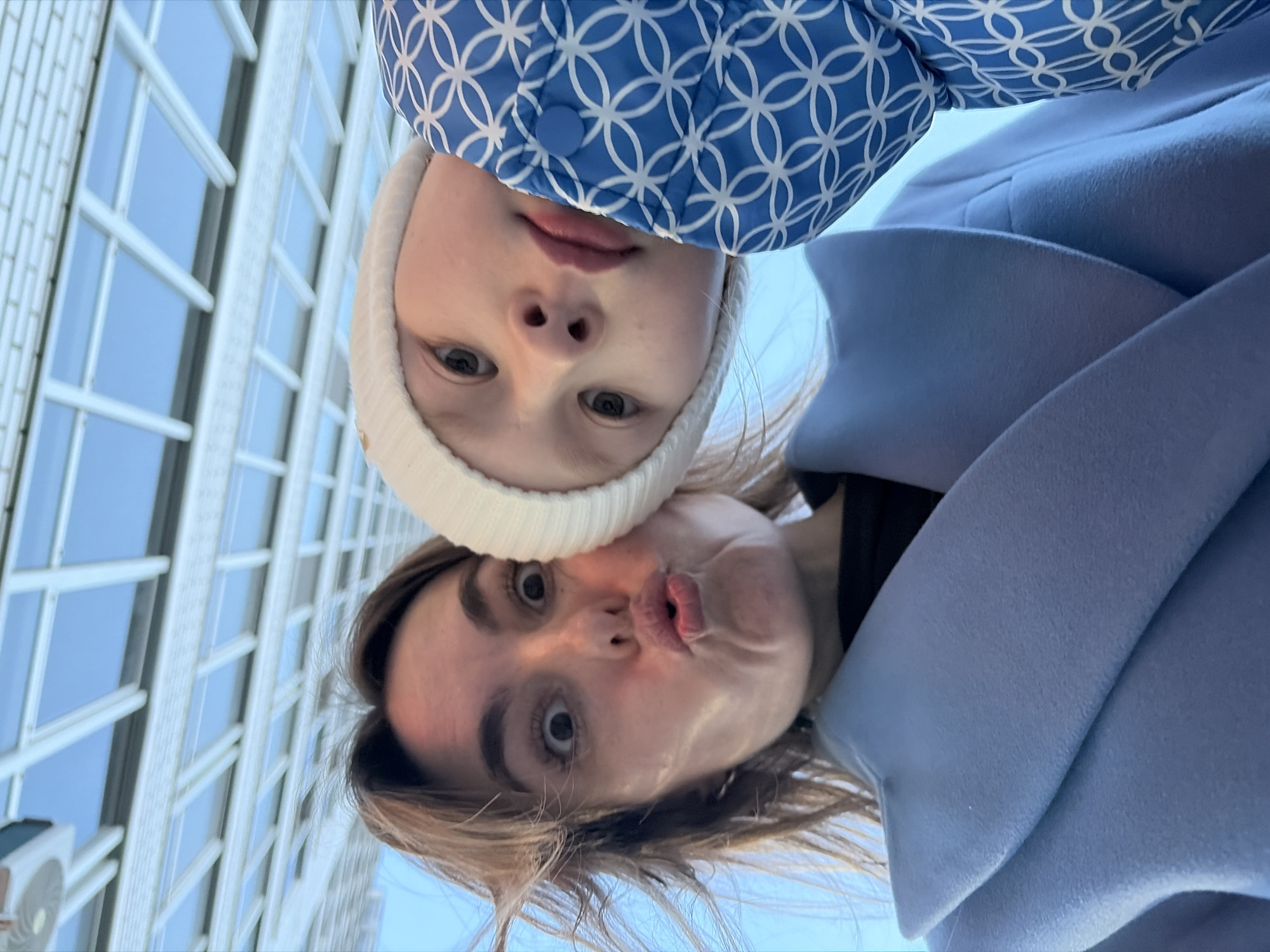 A low-angle, close-up photograph depicts an adult female and a young child, engaged in a selfie against a distinctive architectural ceiling. The adult female, positioned on the left, has light brown hair, wide-open eyes, and pursed lips, making a playful "fish face" expression. She wears a light periwinkle-blue overcoat with broad lapels over a dark garment. To her right, a young child is partially visible, leaning into the frame with a neutral expression, looking directly at the camera. The child wears a white knit beanie and a blue puffer jacket with a repeating white geometric (interlocking circle) pattern and a blue snap button. The background features a modern indoor ceiling structure composed of white structural beams forming a grid, beneath which are numerous light-blue, translucent rectangular panels, suggesting skylights. This architectural feature identifies the location as the **Shopping Center Talisman (ТРЦ Талисман)** in Izhevsk, Russia.