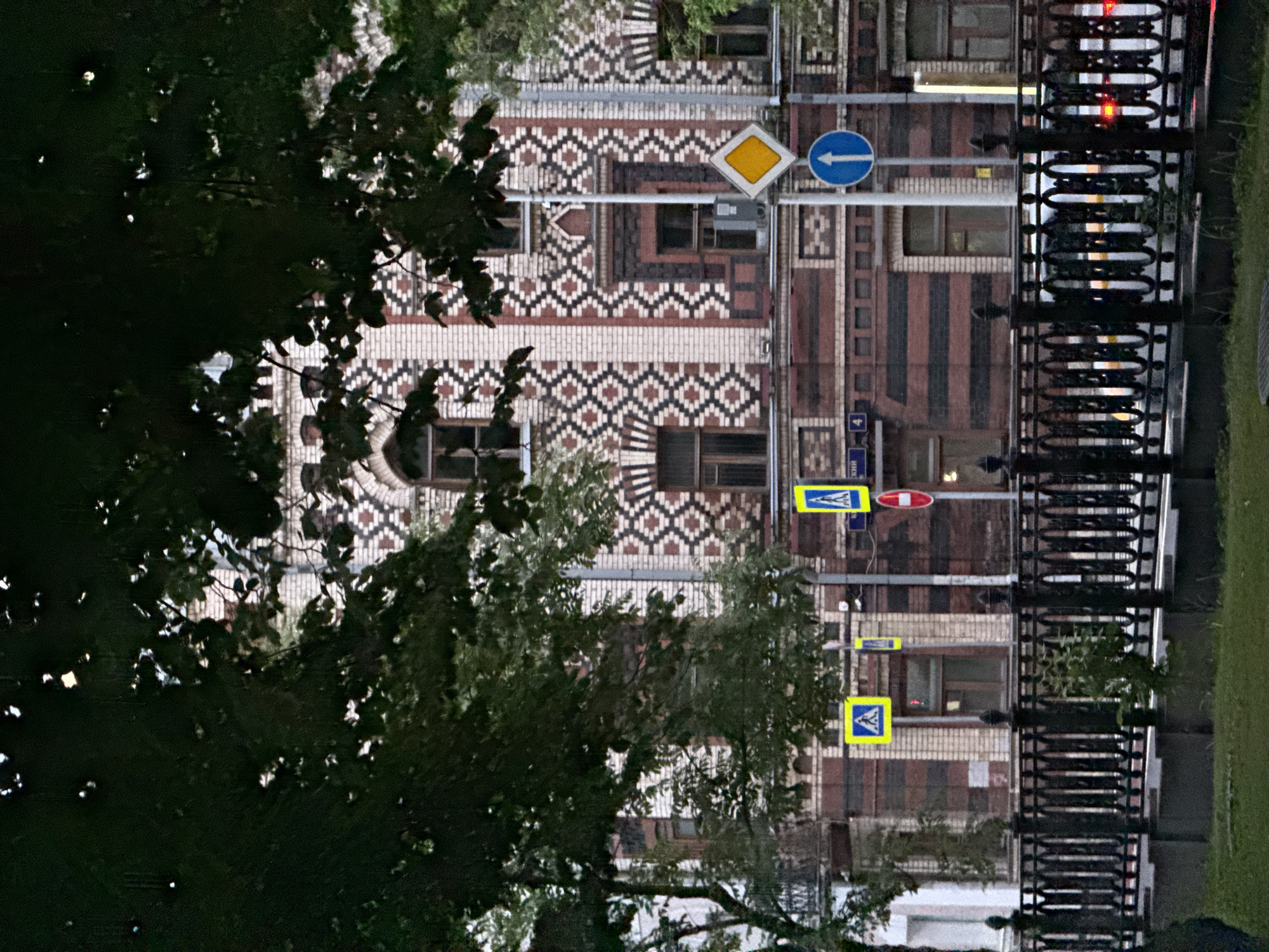 This is an outdoor, eye-level shot of a building in Moscow, Russia. The building has intricate brickwork featuring geometric patterns in varying shades of brown, white, and black. There are windows visible in the building.
Several street signs are present, including a yellow diamond-shaped sign, a blue circular sign with a white arrow pointing left, and rectangular pedestrian crossing signs.
A wrought-iron fence is also visible along the foreground.
Trees and foliage partly obstruct the view of the building.