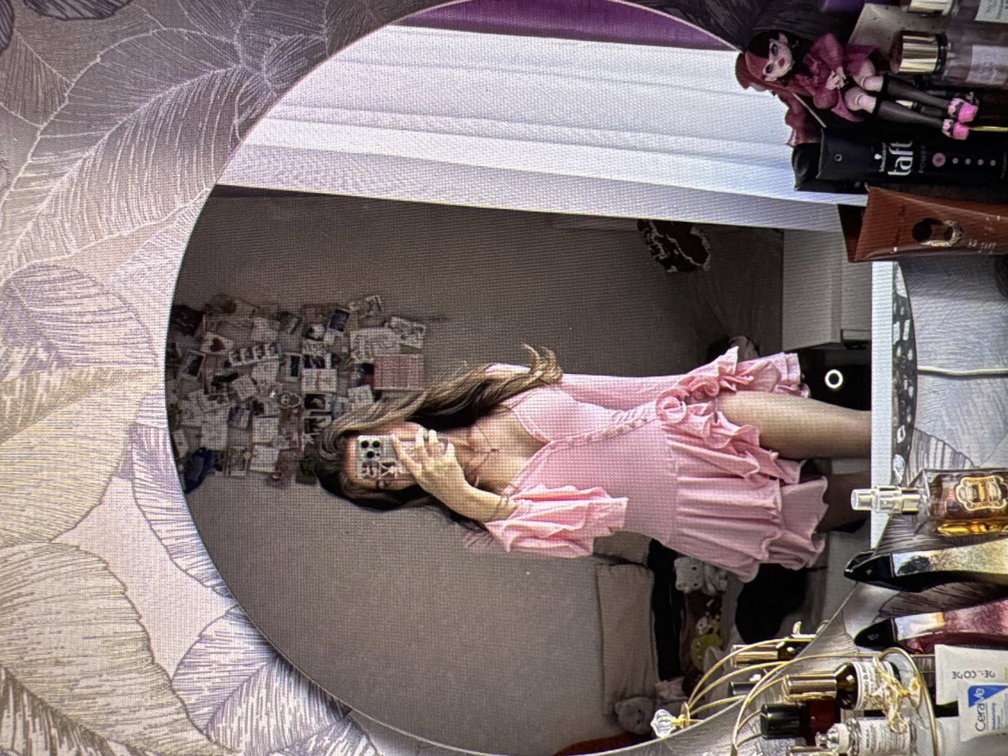 A person is reflected in a mirror, holding a phone to take a selfie. The person is wearing a pink dress with ruffled sleeves and hem. The background shows a bedroom with a bed, a wall decorated with photos, and a shelf filled with various bottles and containers. A doll is placed on the shelf. The mirror is framed by wallpaper with a leaf pattern. The scene is indoors. There is no indication of the city of Castelo Branco, Portugal.