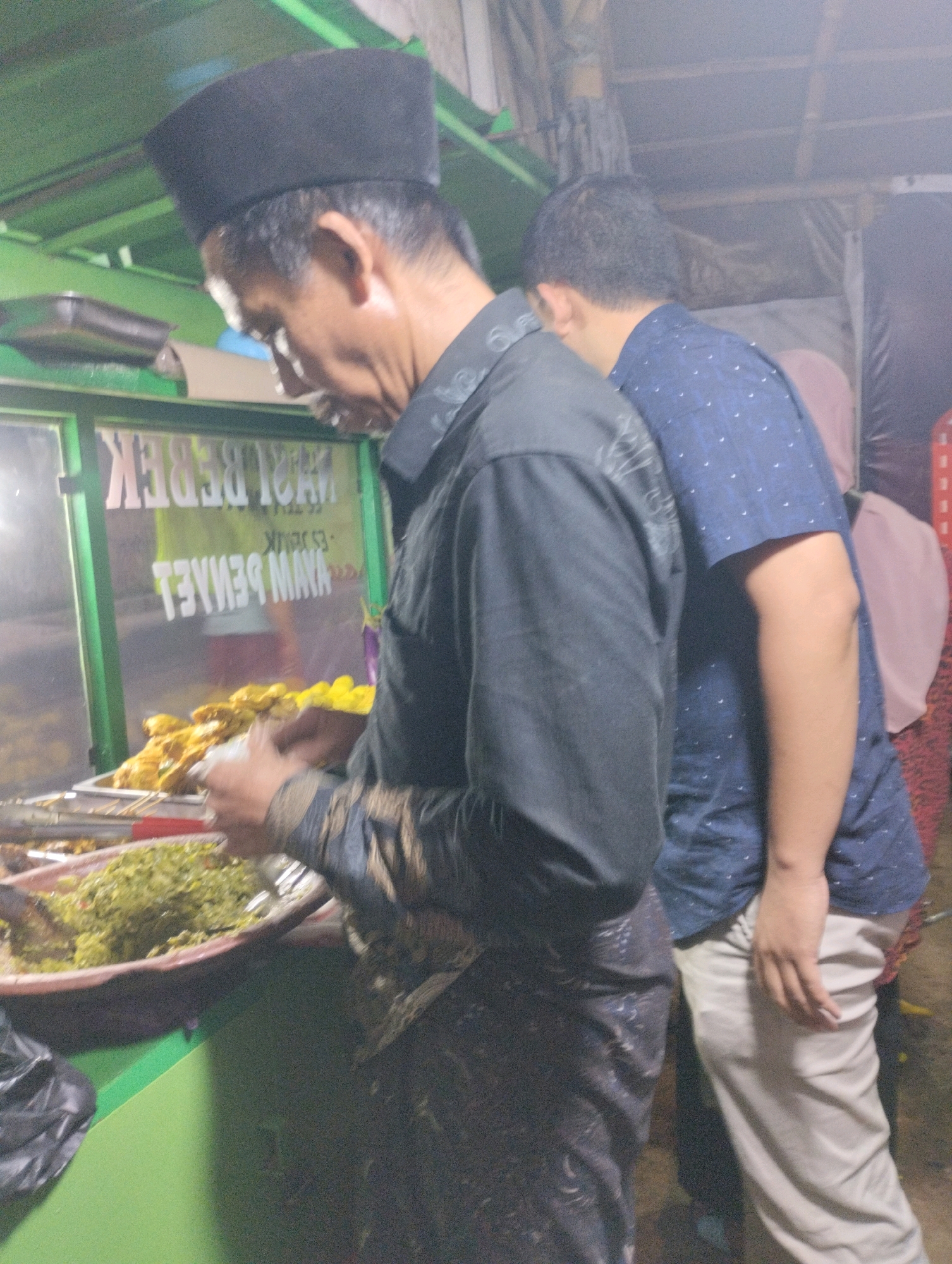 The image captures a street-level food stall in Bekasi, Indonesia, likely operating in the evening given the artificial lighting. In the foreground, a man wearing a black peci (a traditional cap) and a black patterned shirt is partially visible from the side. He appears to be serving or arranging food from a display case. His left hand is holding a white object, possibly a container or a piece of food. Behind him and to his right, another person in a blue patterned shirt and light-colored pants is also partially visible, facing away from the camera.
The food stall's glass display case is illuminated, revealing trays of food. One tray prominently features cooked items, possibly fried chicken or similar dishes, arranged on skewers. Another tray contains a green, finely chopped dish, likely a vegetable side or sambal. The stall's signage is visible on the glass, with text that appears to be in Indonesian, including "AYAM PENYET MAYA" and "MAKI BEBEK" (though reversed due to reflection). The stall itself has a green counter. The overall environment suggests a bustling, outdoor food vending area common in urban settings.