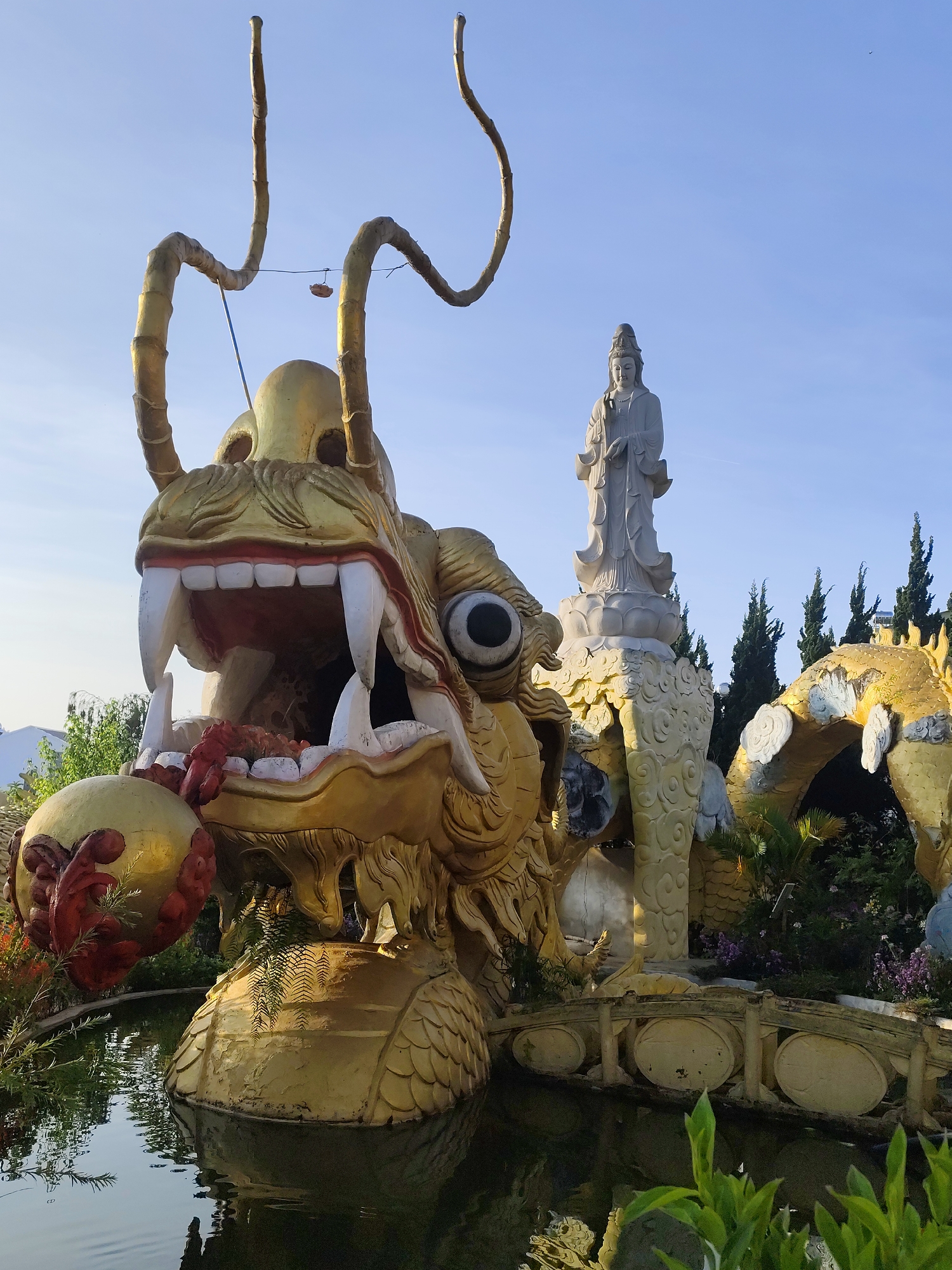 The image depicts a golden dragon statue at a temple in Cam Ly - Đà Lạt, Vietnam. The dragon's head is prominent, with an open mouth displaying teeth and holding a red and gold orb. Behind the dragon is a statue of a standing figure on an elevated, ornate platform with an archway. A small bridge spans a body of water at the base of the dragon statue. Trees are visible in the background, and the sky is light blue. The scene is outdoors.