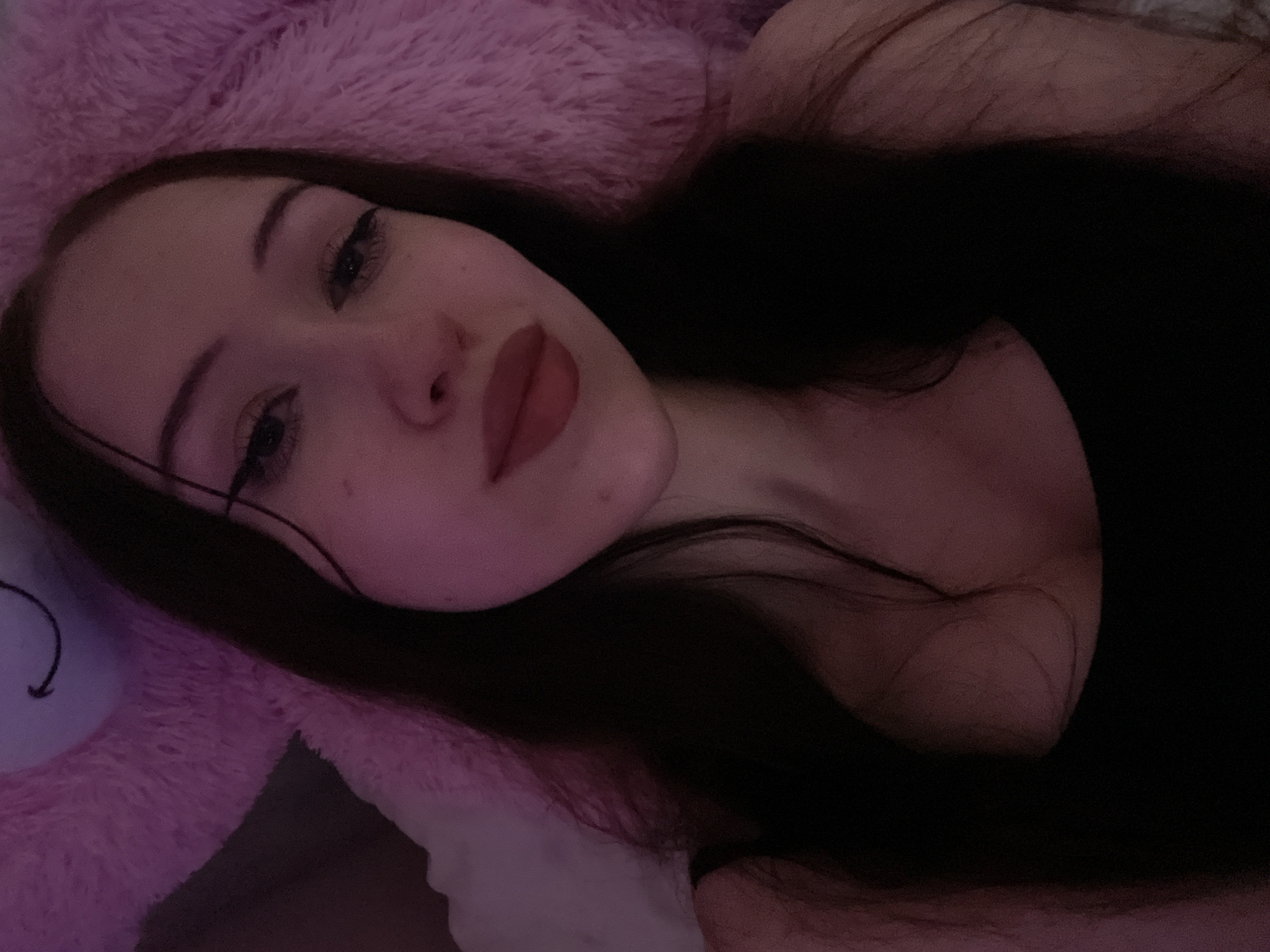 A young woman with long, dark hair is shown in a close-up, selfie-style photograph. She is wearing a black tank top. Her face is turned slightly towards the camera, and she is looking directly at it. She has dark eyebrows, eyeliner, and dark lipstick. Her skin appears pale, and there are some freckles visible on her cheeks. She is lying down, as indicated by the soft, fuzzy pink material behind her head, which appears to be a large stuffed animal or blanket. The lighting in the image is dim, with a pinkish hue, suggesting it might be nighttime or a room with colored lighting. The background is mostly out of focus, drawing attention to the woman's face.
The location in Votkinsk, Russia cannot be determined from the image itself.