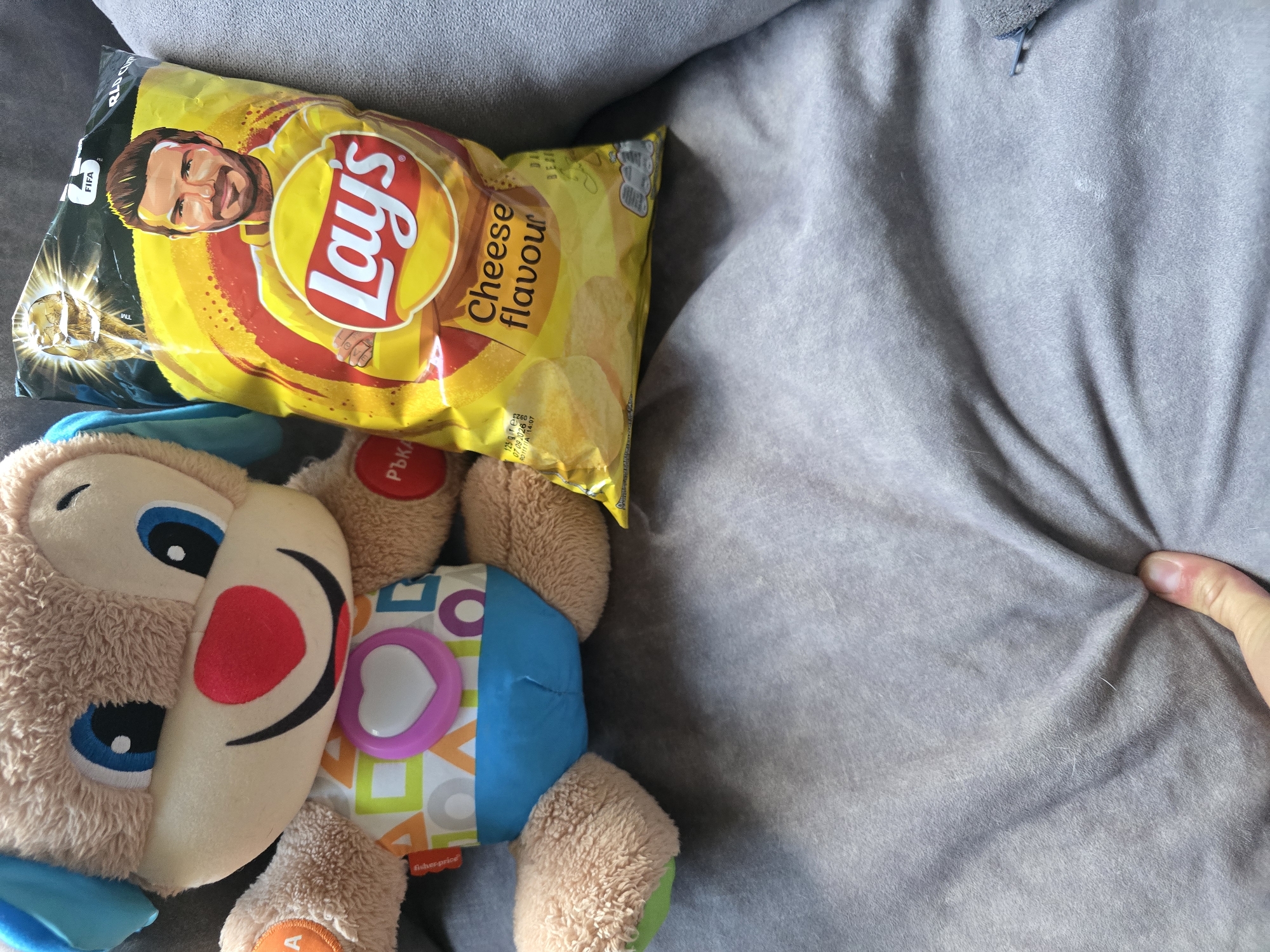 A close-up, top-down shot shows a stuffed toy dog and a bag of Lay's potato chips on a gray, textured surface, possibly a sofa. The stuffed toy, a Fisher-Price Laugh & Learn character, is light brown with blue ears, a red nose, and a colorful, geometric-patterned shirt with a pink heart on it. To the right of the toy is a yellow bag of Lay's chips featuring a picture of David Beckham. The bag is open at the top, and the text "Cheese flavour" is visible. The surface is wrinkled and creased, suggesting it is a soft material. The lighting is bright, casting some shadows. The location in Golyanovtsi, Bulgaria, is not discernible from the image itself.