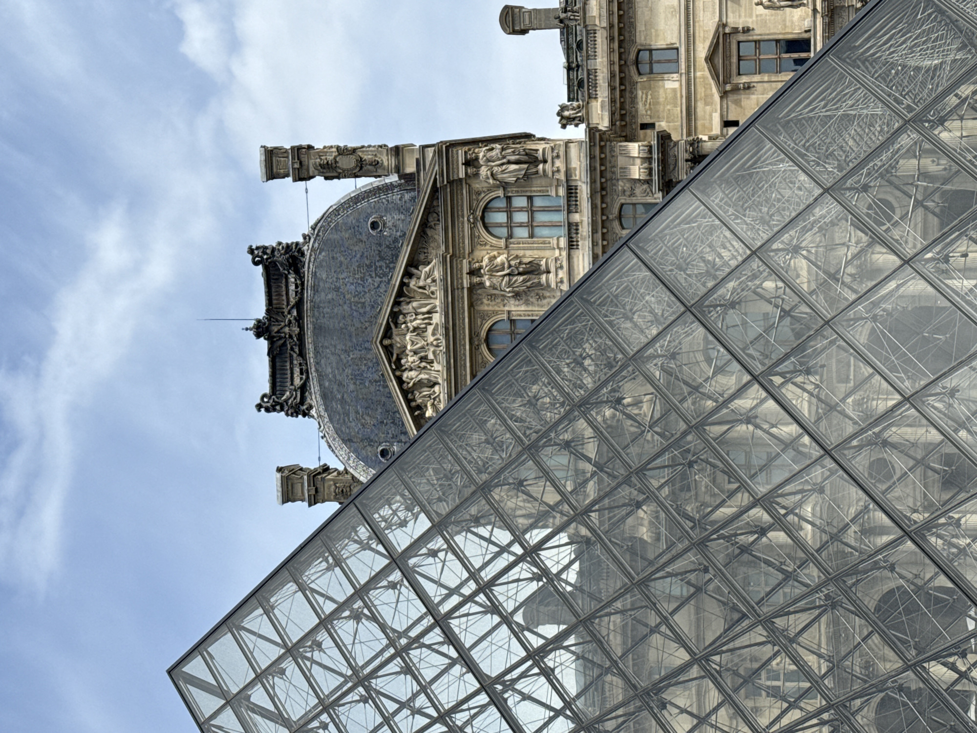 The image captures a view of the Louvre Museum in Paris, France, focusing on the glass pyramid structure and a portion of the historic palace behind it. The pyramid's geometric pattern is prominent, with a grid of glass panels and metal framework. The palace section visible features classical architecture with ornate details, sculptures, and windows. The sky above is pale blue with some cloud cover.