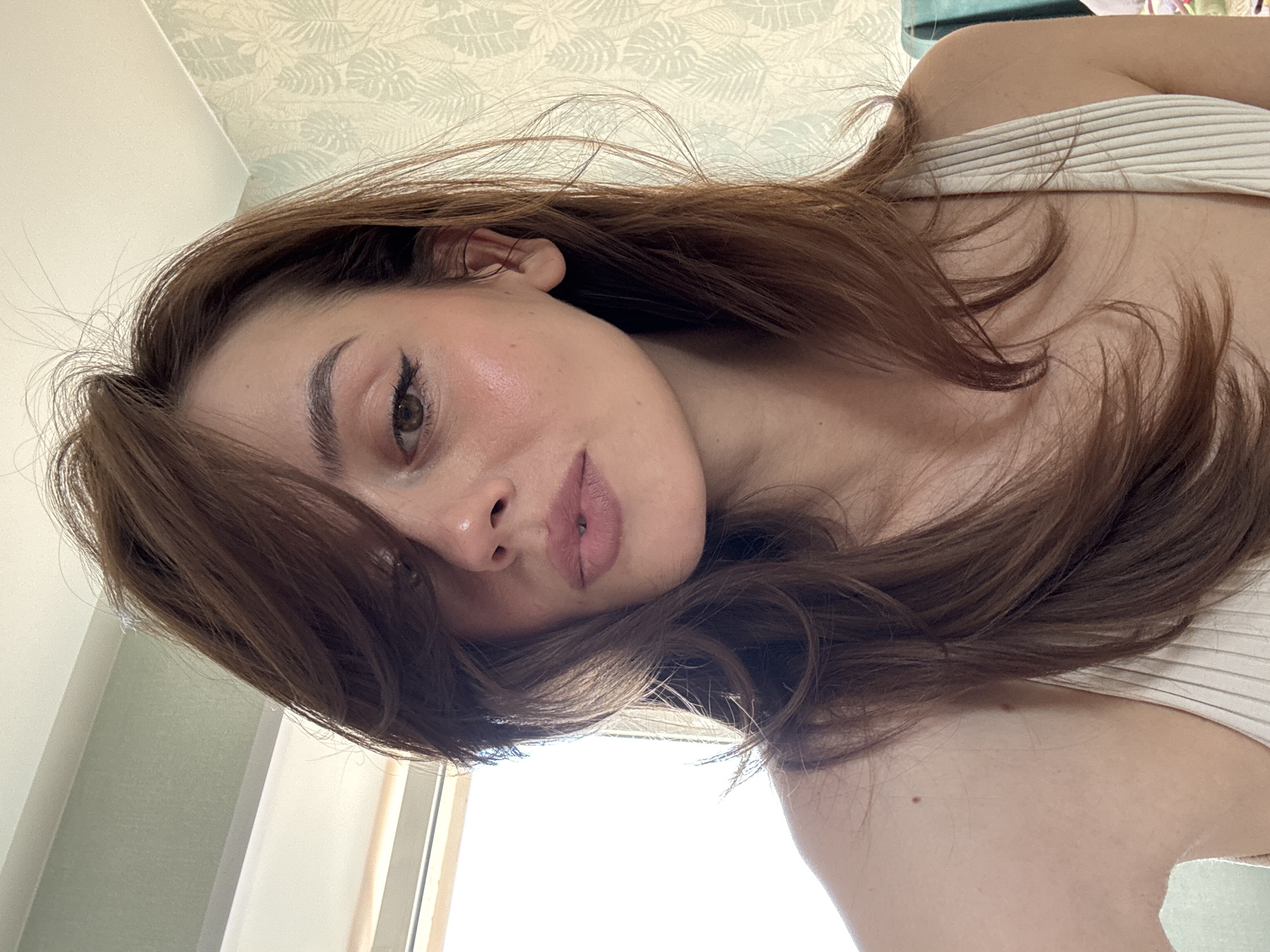 The image, taken in Krasnoyarsk, Russia, features a close-up selfie of a young woman with long, wavy reddish-brown hair. The woman's head is tilted, and she looks directly at the camera with a calm expression. She has visible makeup, including winged eyeliner and a muted pink lip color, and her skin exhibits a natural sheen or blush on her cheeks.
She is wearing a light-colored, possibly cream or beige, ribbed top or tank top. Her long hair frames her face and flows over her shoulders. Small moles are visible on her shoulder and neck area.
The background shows an indoor setting, brightly lit by natural light streaming in from a window located on the right side of the frame. Behind her, a wall is visible, adorned with subtle, light-toned botanical or leafy patterned wallpaper. A ceiling corner is also present in the upper left of the image. The bright illumination suggests the photo was taken during the daytime. No text is visible in the image.