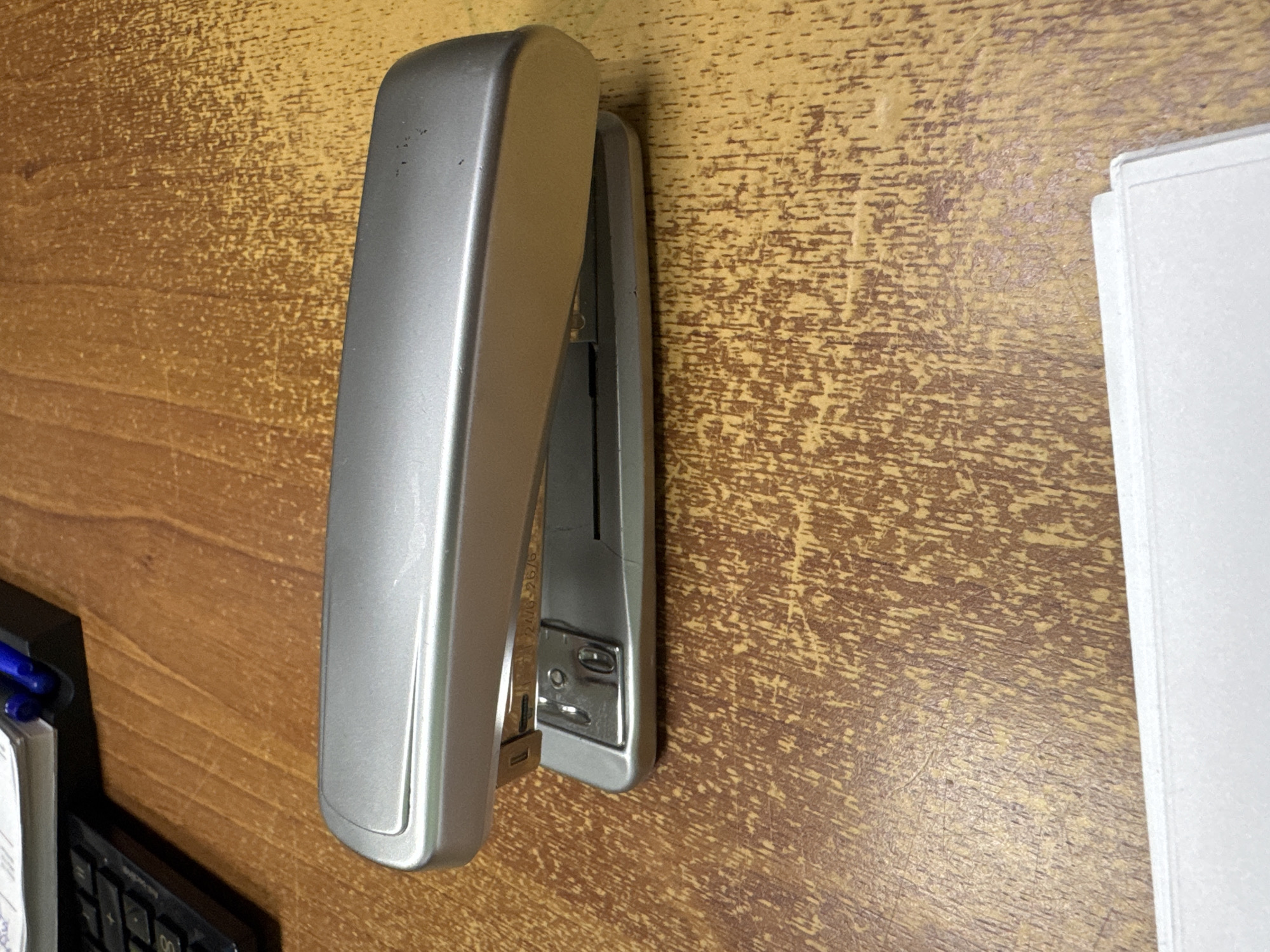 A silver stapler is positioned diagonally on a wooden desk. Part of a calculator and some pens are visible in the top left corner, along with a stack of papers behind them. The desk surface is textured with wood grain. The bottom of the image shows a portion of a white paper or binder. This scene is not depicted in the city of Pyatigorsk, Russia.