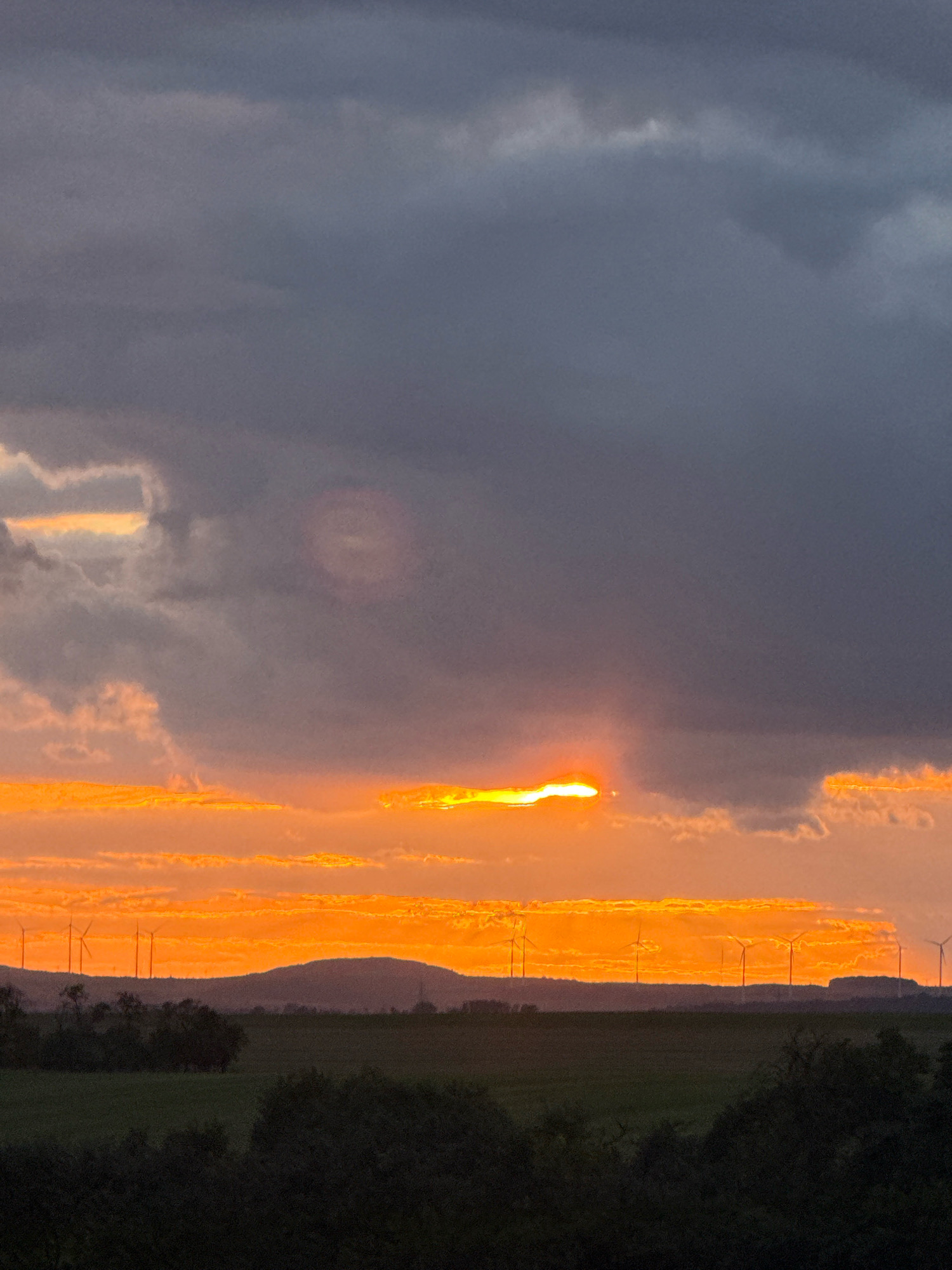 This image captures a dramatic sunset over a rural landscape in Greußen, Germany. The sky is dominated by dark, heavy clouds in the upper portion, with a band of brilliant orange and yellow light breaking through near the horizon. The sun itself is partially obscured by a thin layer of clouds, creating a bright, intense glow.
In the middle ground, a silhouetted line of hills stretches across the frame, with several wind turbines visible atop them. The wind turbines are tall and slender, their blades appearing stationary against the dramatic sky. Below the hills, a dark green field is visible, suggesting agricultural land.
The foreground is filled with the dark silhouettes of trees and shrubs, adding depth to the scene and framing the more brightly lit middle and background. The overall impression is one of a powerful natural event occurring in a quiet, sparsely populated area. There are no people or animals visible in the image. The scene appears to be a landscape photograph taken during the late evening.
