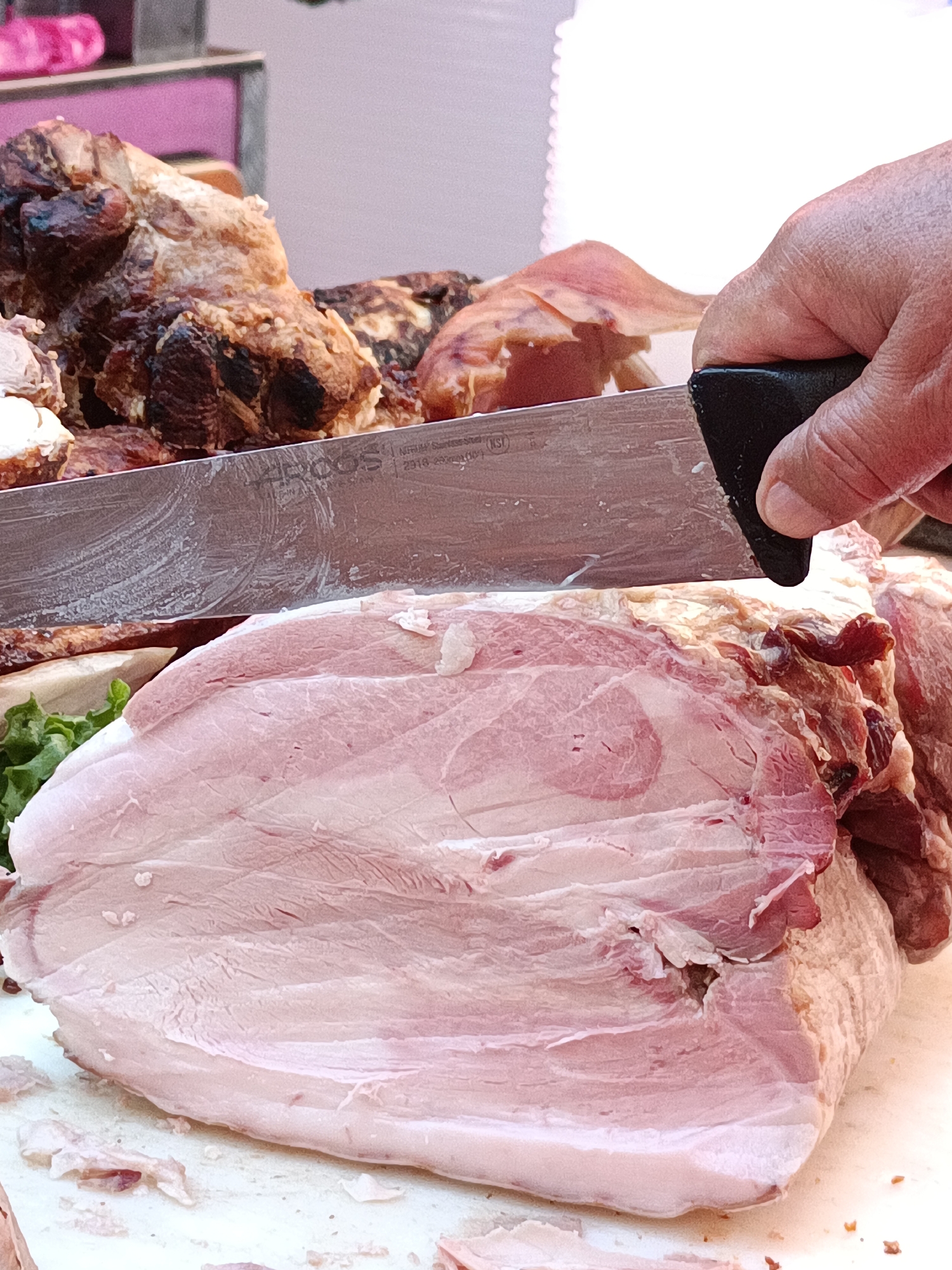 A person's hand grips a large knife, held horizontally, as it slices into a substantial cut of raw pork. The pork is a pale pink with distinct layers of fat and muscle, some darker, reddish areas, and white rind on top. In the background, there are other pieces of cooked meat, appearing roasted or smoked, with a dark, crusted exterior. The scene suggests a food preparation or market setting. The image is a close-up, focusing on the action of slicing the meat.