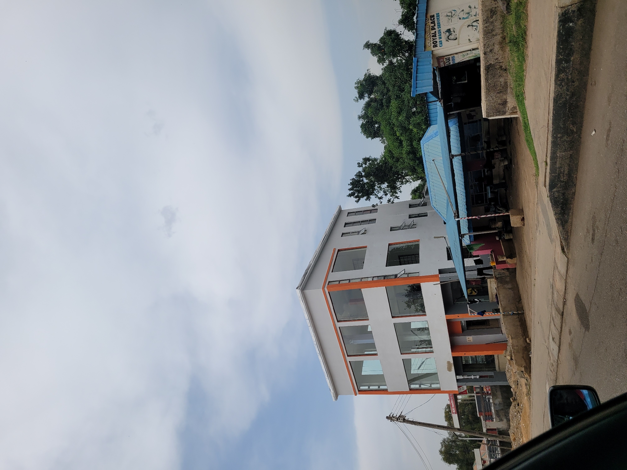 The image shows a three-story building in Gura Top, Nigeria. The building is white with orange trim around the windows and edges. It has large windows on the front facade. The building is situated next to a smaller structure with a blue roof that houses "Royal Place Car Wash Services." A street and sidewalk are visible in the foreground. Utility poles and wires are also visible, as is part of a vehicle's side mirror in the lower right corner. The sky is mostly overcast with some patches of blue.