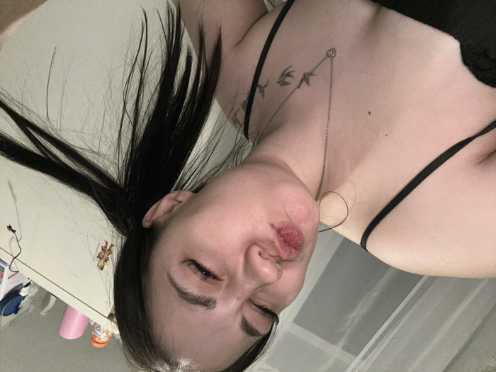 The image depicts a female individual indoors, likely within a residential setting in Chaikovsky, Russia. The specific location within the city cannot be determined from the visual evidence. The subject, a female with dark hair styled in pigtails or ponytails, occupies the majority of the frame. Her face is turned slightly to the left, with her eyes partially closed and her lips puckered in a "kissing" expression. She wears a black top with spaghetti straps and a silver-colored chain necklace featuring a heart-shaped pendant. A tattoo of three bird silhouettes is visible on her left collarbone/shoulder area. The background features a light-colored curtain or sheer fabric on the left. On the right, a white cabinet or wardrobe is visible, with several objects on its top surface, including an orange container, a pink cylindrical object, a blue object, and a black electrical cord. A small figure, appearing to be a decorative item in a red outfit, is affixed to the cabinet door.