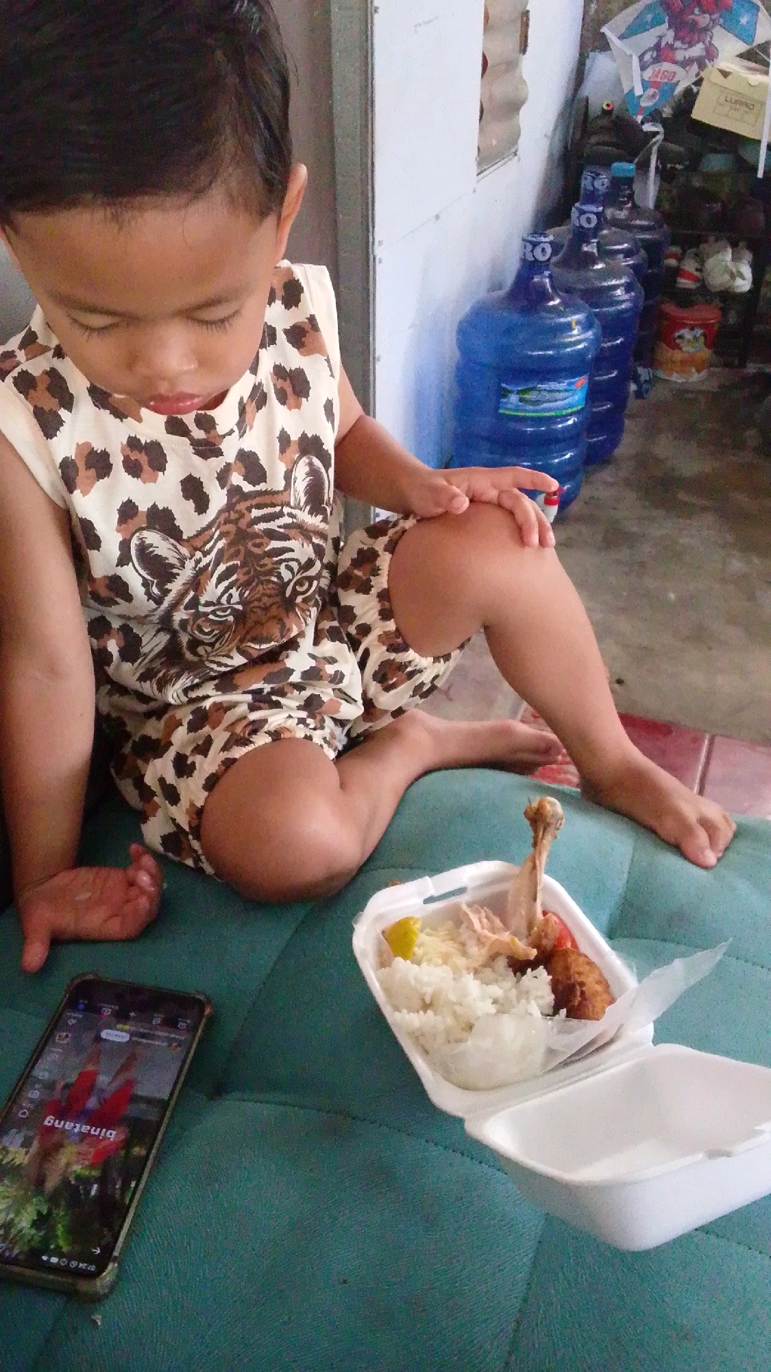 A young child, wearing a sleeveless shirt and shorts with a leopard print and tiger graphic, sits on a teal-colored cushioned surface. The child is looking down at a white styrofoam container filled with rice, chicken, and what appears to be a tomato and lemon slice. To the left of the child is a smartphone displaying a video with text that reads "binatang". In the background, several large blue water jugs are lined up against a wall, and various items, including a kite, are visible on shelves. The scene appears to be indoors, possibly in a home or a public area with basic furnishings. The lighting suggests daylight.