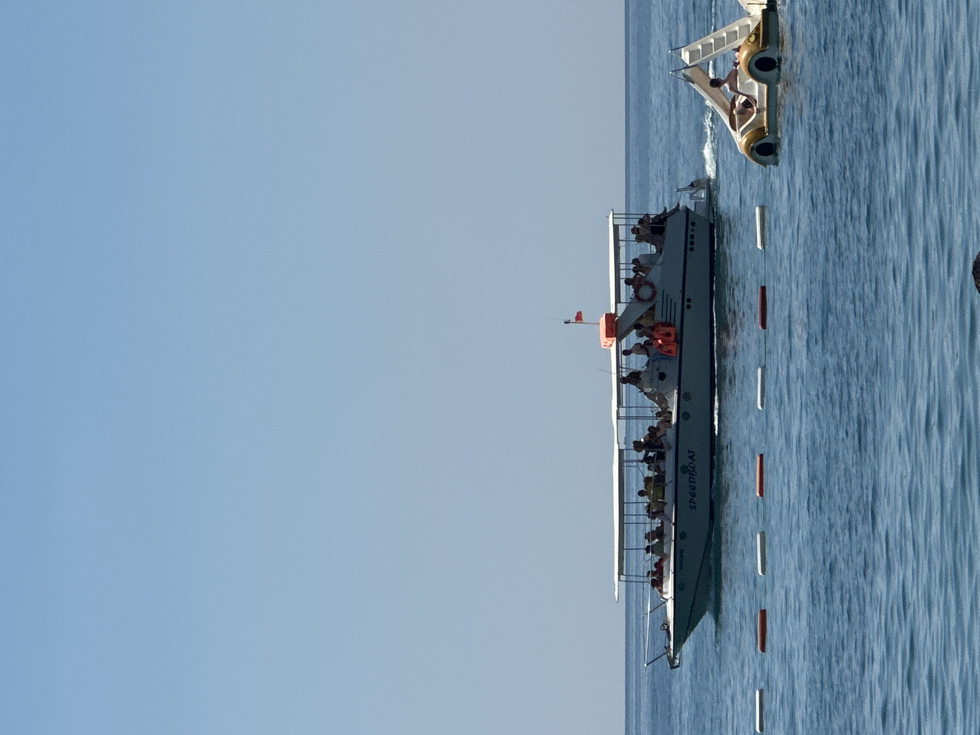 In Boreti, Montenegro, a large speedboat carrying numerous passengers cruises across a calm, blue sea under a clear sky. The boat, with a white canopy providing shade, has "SPEEDBOAT" written on its side. In the foreground, a smaller, yellow, car-shaped pedal boat with a single person steering it is visible. A line of buoys, alternating between white and orange, stretches across the water between the two vessels. The overall scene depicts a typical sunny day at a coastal resort.