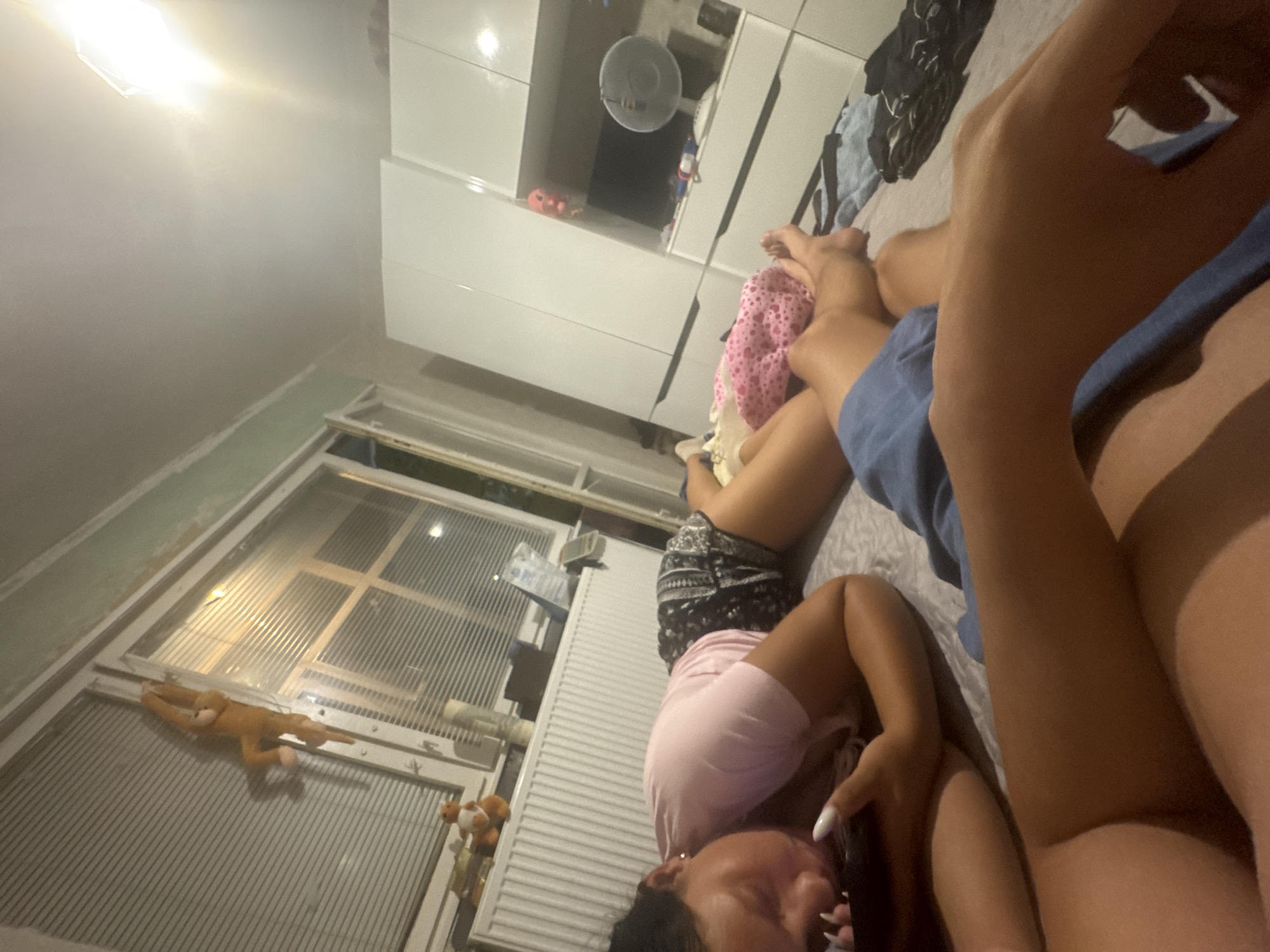 The image depicts a bedroom scene. In the foreground, two individuals are lying on a bed. One person, in the lower right, is wearing blue shorts and their legs are visible. Another person, in the mid-left, is lying on their side, wearing a pink t-shirt and patterned shorts, and appears to be using a mobile phone. A pink and white spotted blanket is visible near their legs.
In the background, there is a large white wardrobe with multiple doors. To the left of the wardrobe is a television set and a small white fan on a shelf. Above the television, there are some items on a shelf, including what appears to be a pink stuffed animal. To the left of this, there is a radiator and a window with horizontal blinds. A plush toy monkey is hanging from the window frame. The overall lighting suggests it is either evening or night, with a bright light source visible in the upper right corner.
While the image captures elements of a domestic setting, there is no visual information within the image to indicate the location is the city of Sofia, Bulgaria.