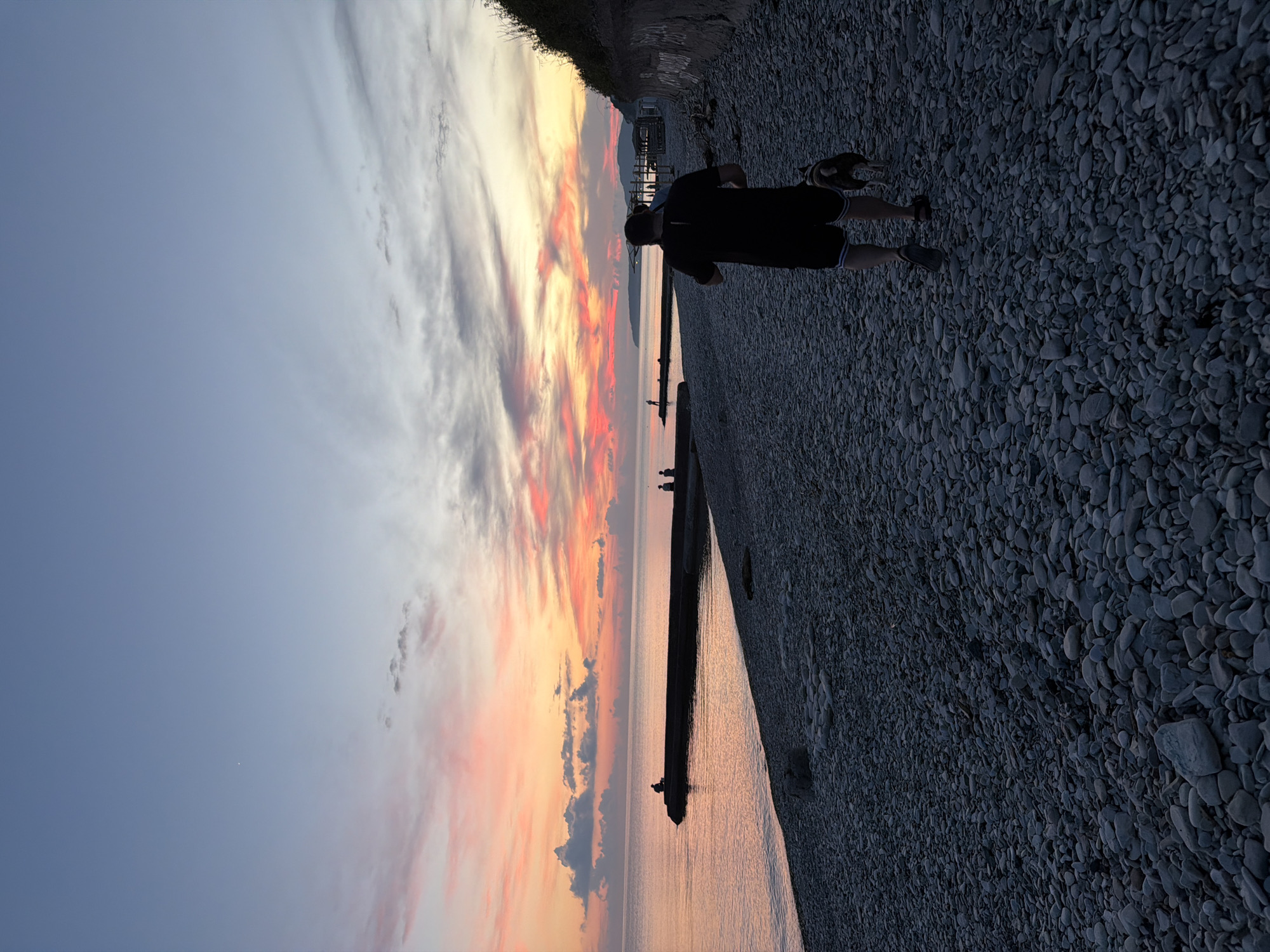 A man walks his dog along a pebble beach at sunset. The sky is a gradient of blue, pink, and orange, with some wispy clouds. The ocean is calm and reflects the colors of the sky. In the distance, a pier extends into the water, with a few people sitting on it. The beach is covered in smooth, gray pebbles. The overall scene is peaceful and serene, evoking a sense of calm and tranquility. The location is the city of Dederkoy, Russia.