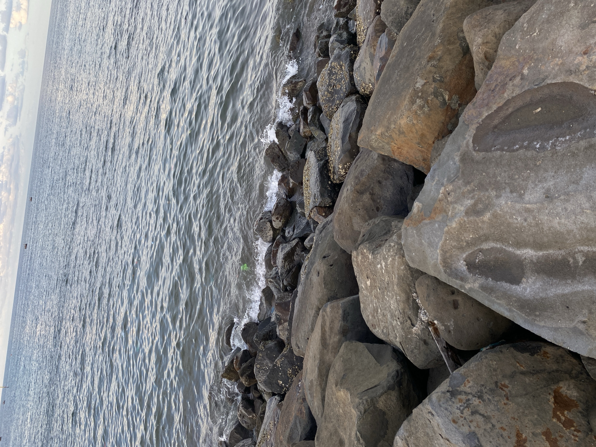 The image depicts a coastal scene in Lhokseumawe, Indonesia. The composition features a body of water occupying the left half of the frame, with gentle ripples on its surface. The right half is dominated by a collection of large, gray rocks forming a breakwater or shoreline fortification. Some of the rocks are encrusted with barnacles. Waves are gently crashing against the rocks. A small piece of green debris is visible near the waterline.