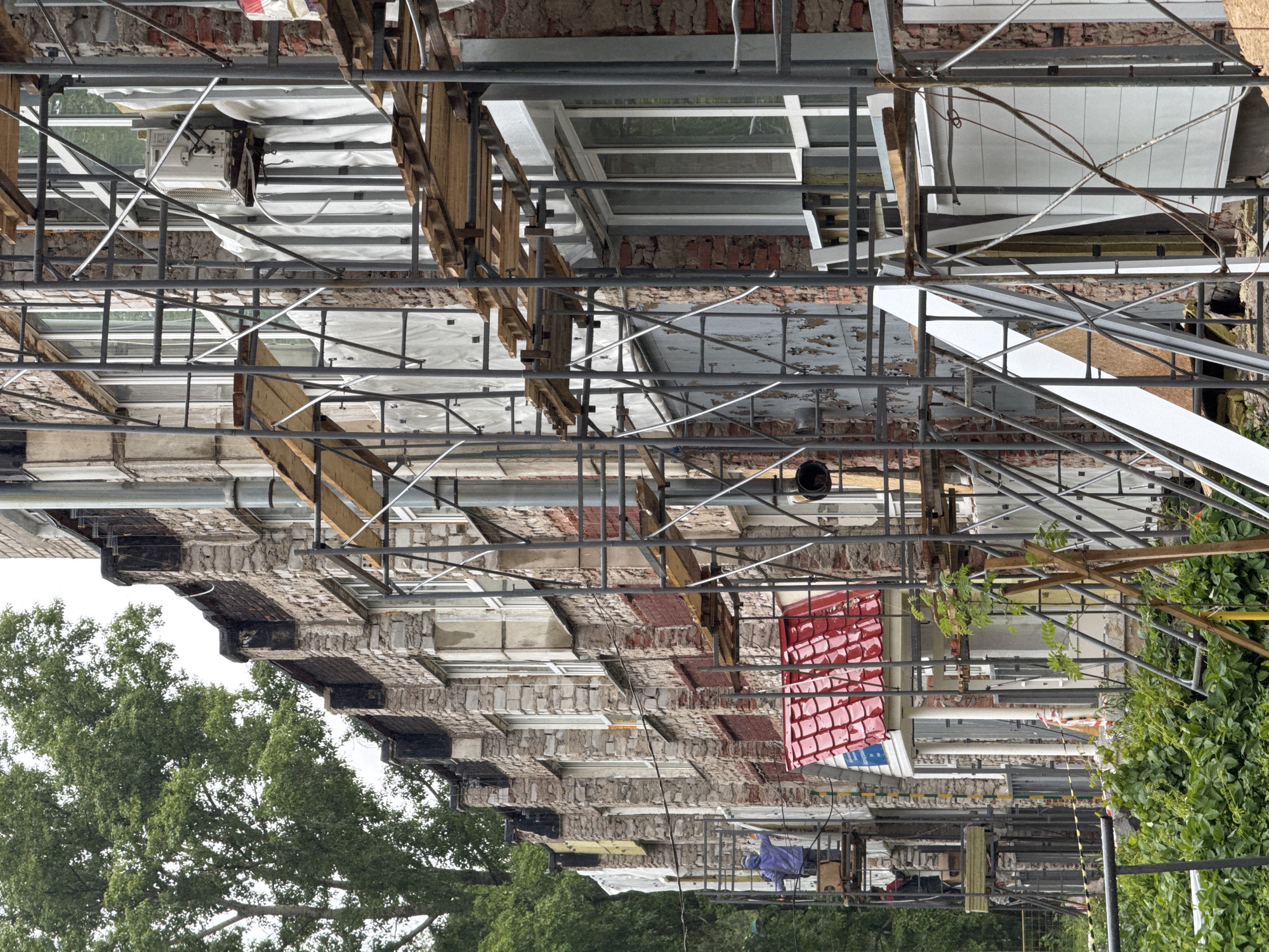 The image captures a multi-story building in Chaikovsky, Russia, undergoing extensive renovation or construction, viewed from a low angle looking upwards. The entire facade of the building is covered by a dense network of metal scaffolding, featuring numerous vertical and horizontal poles, diagonal braces, and wooden planks that serve as walkways for workers and material storage.
The building itself displays exposed brickwork and crumbling plaster in various sections, indicating the historical nature of the structure and the extent of the renovation. Some window openings are fitted with new frames or covered with white protective sheeting, while others remain dark and open. Construction materials such as wooden planks, white insulation panels, and various tools are visible on the scaffolding platforms. An outdoor air conditioning unit is mounted on an upper level, partially covered by white material. On a lower platform, a blue tarp is draped over an object, likely covering equipment or materials.
A section of a new or renovated red-tiled roof is visible towards the right side of the building. In the lower left portion of the image (when oriented correctly), large, leafy green trees are prominent, providing a natural element against the urban construction. The sky appears bright and overcast, indicating daytime conditions without direct sunlight. A small blue sign with white text is visible on the red roof structure, and some caution tape can be seen, but the text on both is unreadable. The scene conveys an active construction site, typical of urban renewal or development.