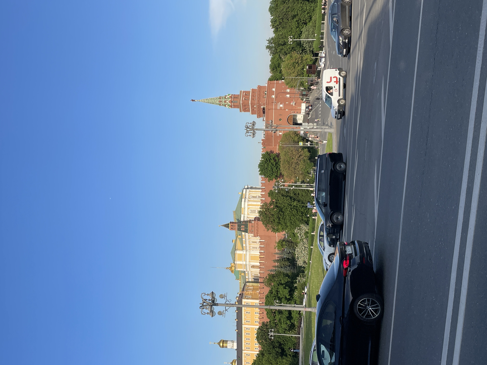 The image shows a view of the Moscow Kremlin from a road. The architecture of the Kremlin, including the brick walls, towers, and golden domes of the cathedrals, is visible. There are trees and green spaces around the Kremlin. Several cars are on the road. The sky is clear blue. The image appears to have been taken from a high vantage point.