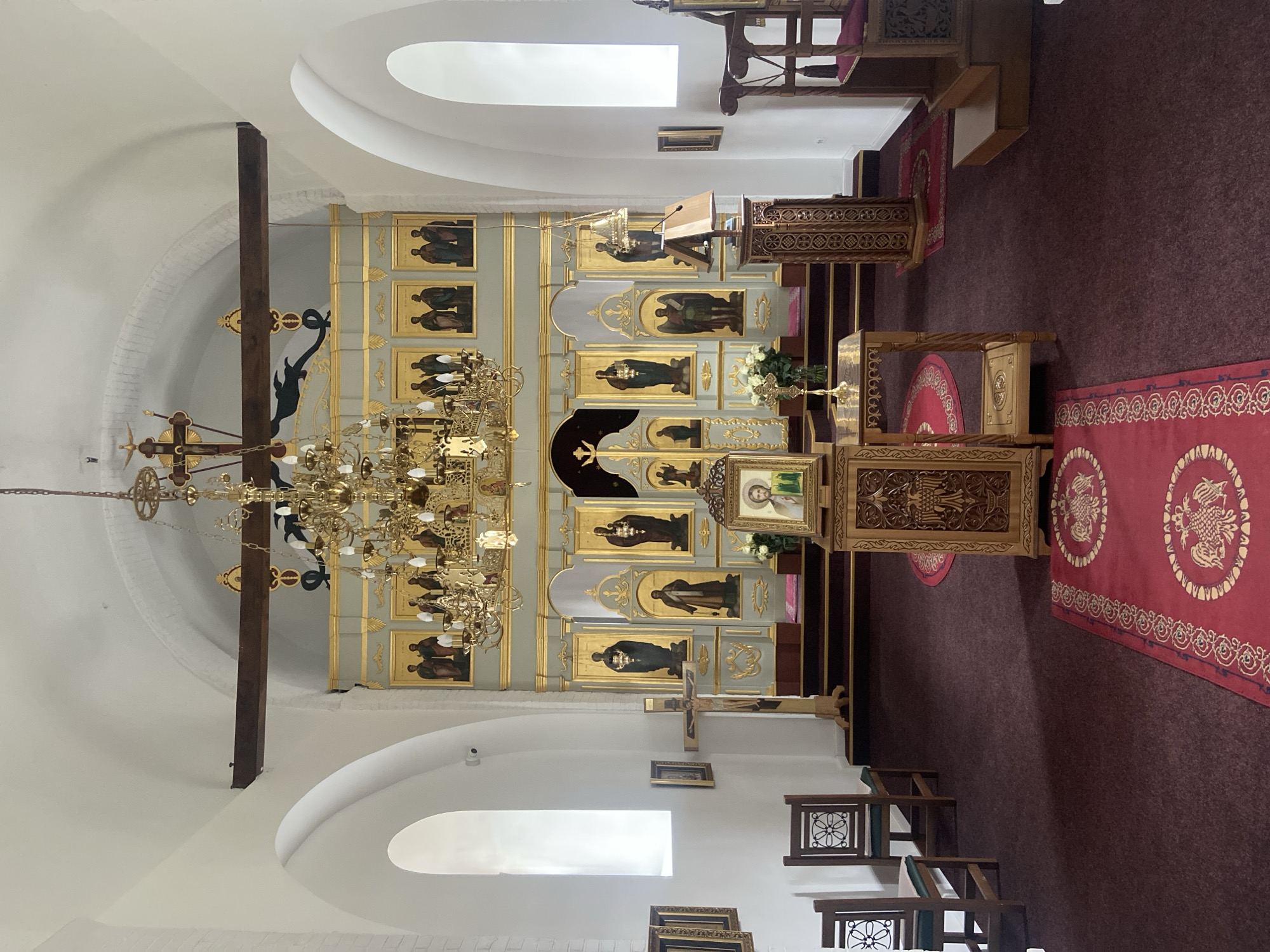 The image depicts the interior of an Orthodox church in Kolašin, Montenegro. The architecture features white-painted walls and arched ceilings. A prominent iconostasis adorned with gold accents and religious paintings dominates the foreground. A large, ornate chandelier hangs from the ceiling. Religious objects like a wooden lectern and a red carpet with golden decorations are visible, implying a place of worship. A wooden cross hangs against the wall. Natural light enters through arched windows. No people are visible.