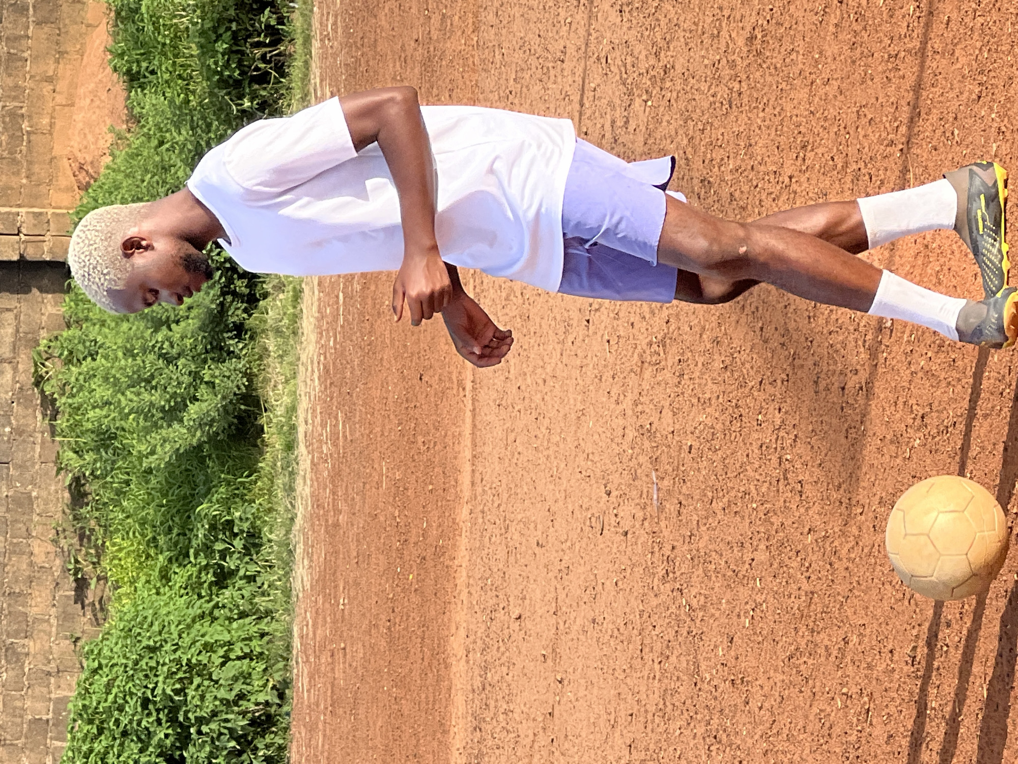 A Black male with bleached blonde hair is standing on a dirt soccer field. He is wearing a white t-shirt, light blue shorts, white socks, and soccer cleats. He is facing to his right, with his left leg crossed in front of his right leg. A light-colored soccer ball is positioned on the ground in front of him, to his left. In the background, there is a brick wall and a dense green bush. The scene is consistent with a soccer field in an urban or peri-urban environment. The quality of the lighting suggests it is daytime, likely late afternoon due to the long shadows.