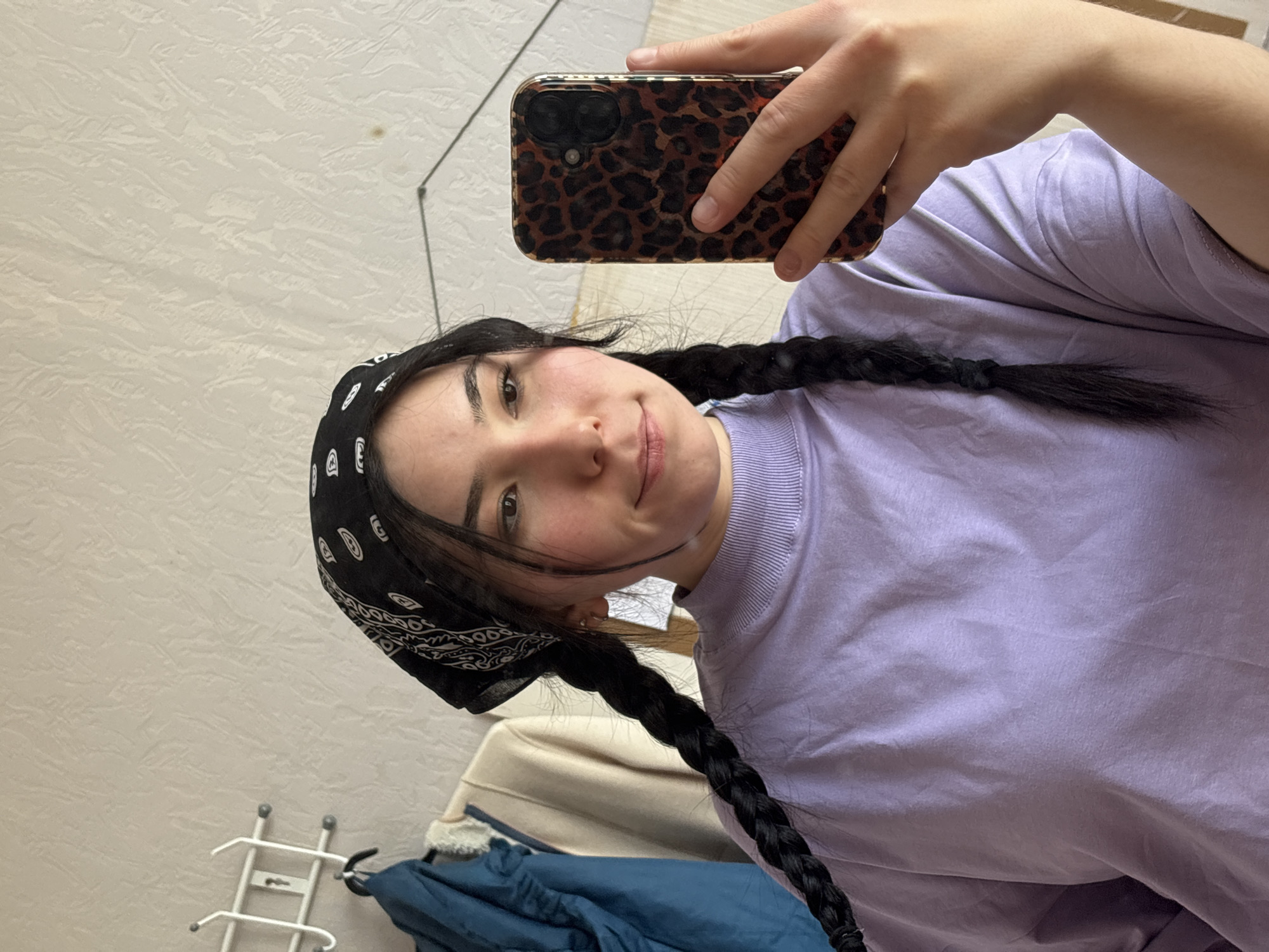 A young woman is taking a selfie in what appears to be an indoor environment. She is wearing a light purple t-shirt and has long, dark braids and a black bandana on her head. She is holding a smartphone with a leopard-print case. Part of a clothing rack with a blue jacket hanging on it is visible in the background. The wall behind her is a neutral color. There is no information in the image to indicate the location is Izhevsk, Russia.