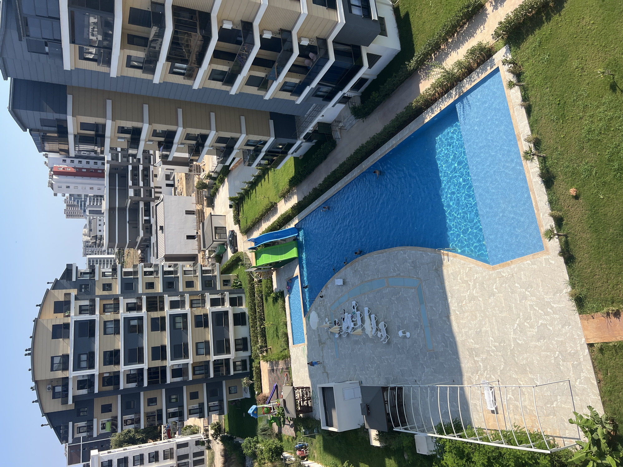 An aerial view shows a residential complex in Aksu, Turkey, featuring modern apartment buildings, a swimming pool, and a children's play area. The buildings are multi-story structures with balconies and contemporary architectural designs. One prominent building is predominantly gray and beige, with dark-framed windows. Another building is white with dark accents. A large, irregularly shaped swimming pool is the central feature of the outdoor space, with sections of varying depths. Several people are visible in the pool, swimming and relaxing. Adjacent to the pool are lounge chairs with umbrellas, a children's playground with slides and swings, and a small gazebo. Lush green grass surrounds the pool area, and pathways made of stone and concrete provide access. The sky is clear and blue, indicating a sunny day.