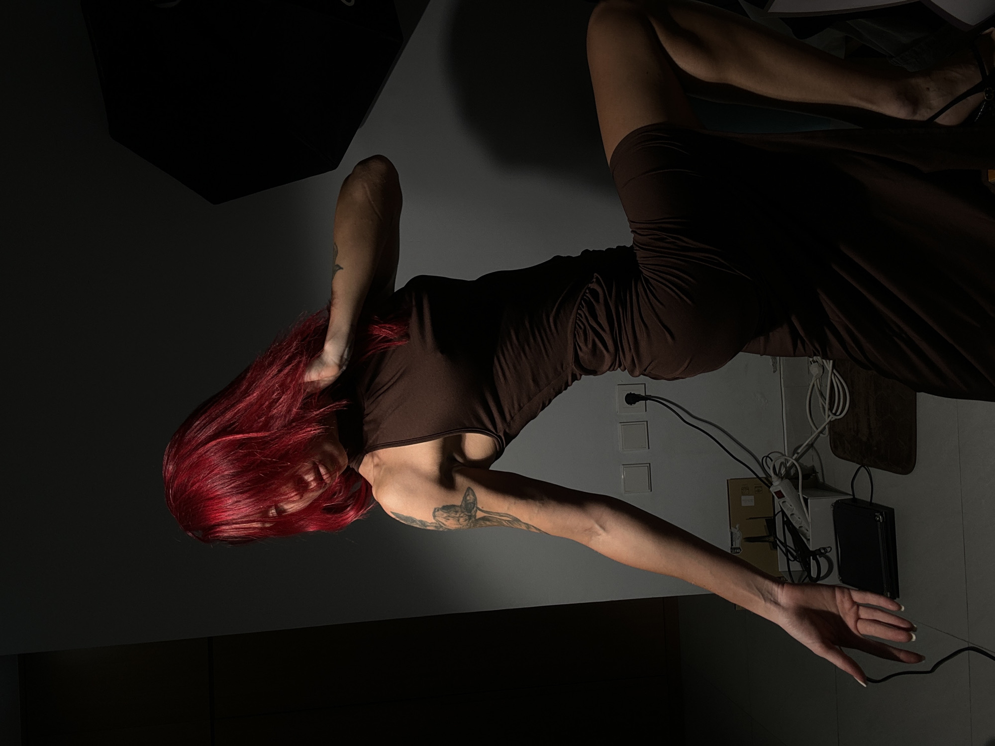 A woman with red hair, wearing a brown dress, is lying on a floor. Her right arm is outstretched and her left arm is bent, with her hand near her face. The background includes electrical outlets, cords, and a black softbox. The location is indeterminate, and there is no information to confirm it is in Dalung, Indonesia.