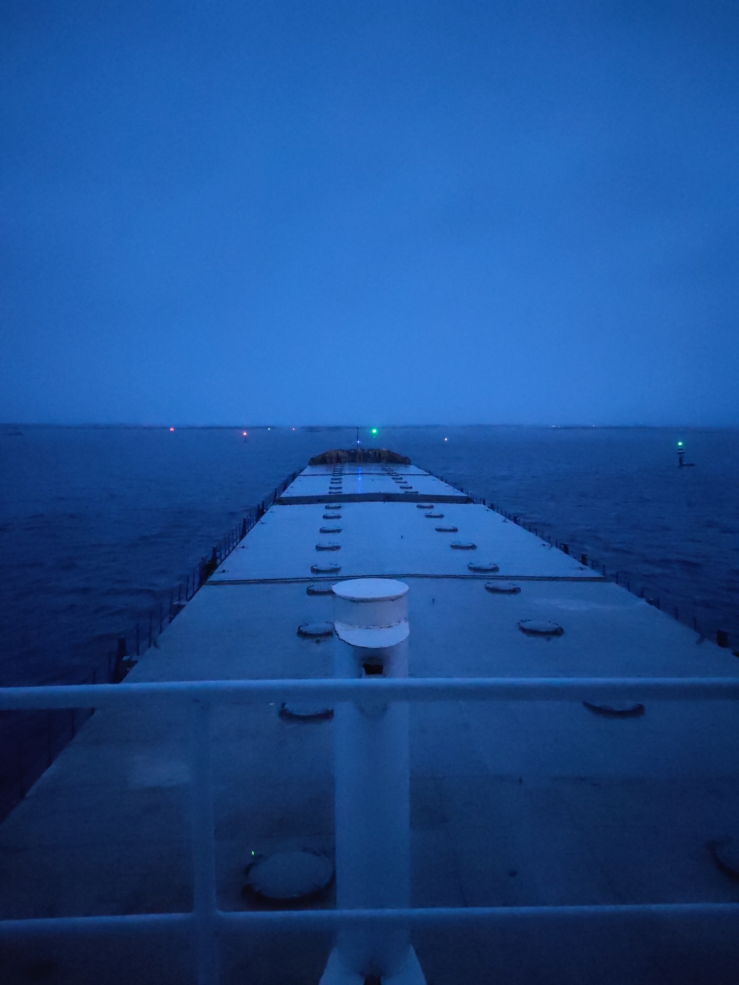 The image depicts a view from the deck of a large ship at night or dusk. The foreground shows a white railing and part of the ship's deck, which appears to be flat and wide with evenly spaced circular hatches. In the distance, the sea stretches to the horizon under a dark, overcast sky. Several lights are visible on the horizon, some green and some red, possibly marking navigational points or other vessels. The overall scene is dimly lit, dominated by shades of blue and grey. No people are visible. The location is not in Bereg, Russia. It is on the open sea.