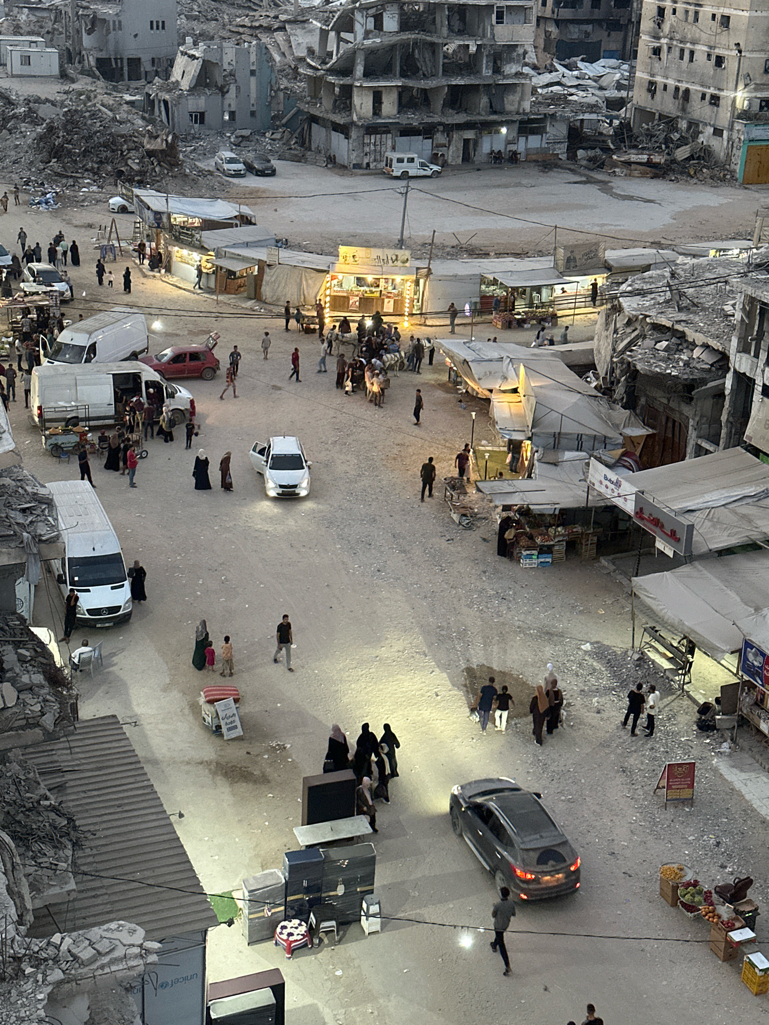 This is a high-angle, long shot of a street scene in Gaza City, Palestinian Territories. The environment is heavily damaged, with destroyed and partially destroyed buildings lining the street. Debris and rubble are visible throughout the scene, suggesting recent conflict. The street is unpaved and dusty.
In the foreground, there are several vans, some with their doors open, and a red car. People are milling about near the vehicles. Further down the street, there are temporary stalls and shops set up, many with tarpaulin coverings, selling various goods, including what appear to be fruits and vegetables.
Several individuals are walking on the street, some in groups, others alone. There are also a few cars on the street, including a white sedan with its door open and a dark SUV with its brake lights illuminated. Some of the buildings appear to be residential, while others look like businesses. The overall impression is one of daily life attempting to resume amidst widespread destruction. The lighting suggests it is either early morning or late evening.