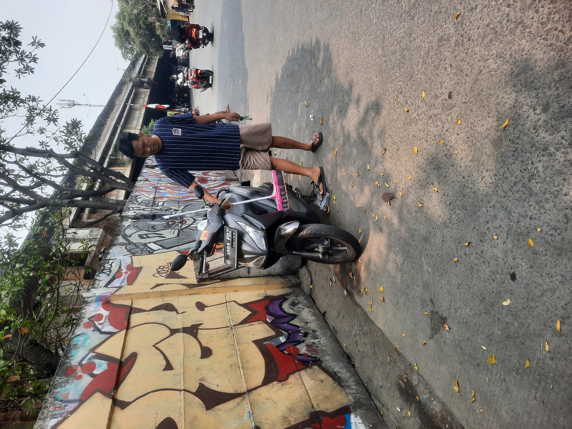 A man stands on a sidewalk next to a parked black scooter. He is wearing a dark blue and white striped t-shirt and light brown shorts, holding keys and a pink-handled broom. The scooter has a license plate that reads "B 3009 PDZ". The sidewalk is paved with concrete and is scattered with fallen yellow leaves. Behind the scooter is a graffiti-covered wall with large, colorful lettering. In the background, a busy street with several other motorcycles and an overpass can be seen, suggesting an urban environment, possibly in North Jakarta, Indonesia, given the license plate format. The lighting indicates it is daytime.