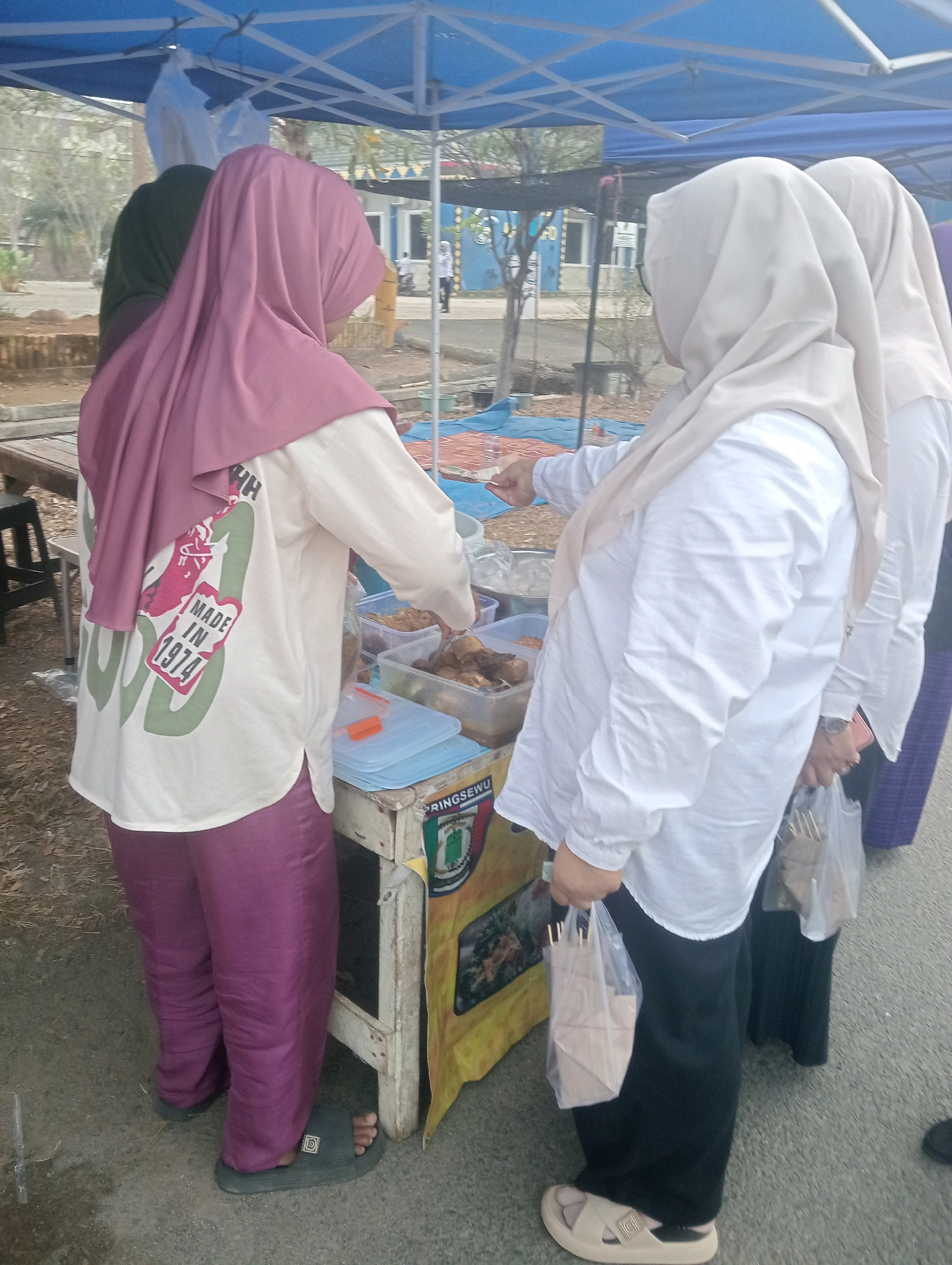This is a vertical, eye-level, full-shot photograph of three women at an outdoor food stall. The women are facing away from the camera and are mostly covered by hijab and modest clothing. The woman on the left wears a light pink hijab and a cream-colored shirt with a graphic print that reads "MADE IN 1974." She also wears purple pants and black sandals. She appears to be looking at the food being sold at the stall. The woman in the middle wears a light beige hijab and a white long-sleeved shirt, with black pants and beige sandals. She is reaching into a clear plastic container filled with food. The woman on the right wears a light beige hijab and a white long-sleeved shirt and a long dark skirt. She is holding two transparent plastic bags, each containing what appear to be food items wrapped in brown paper.
The food stall is set up under a blue canopy. The stall itself is a simple wooden table with a yellow and patterned tarp draped around its front. Various plastic containers of food are visible on the table, along with a blue container with an orange lid. The ground surface is asphalt. In the background, there are trees and buildings, suggesting an urban or semi-urban environment. The overall scene appears to be a market or an outdoor event. The location is identified as Sidoharjo, Indonesia.