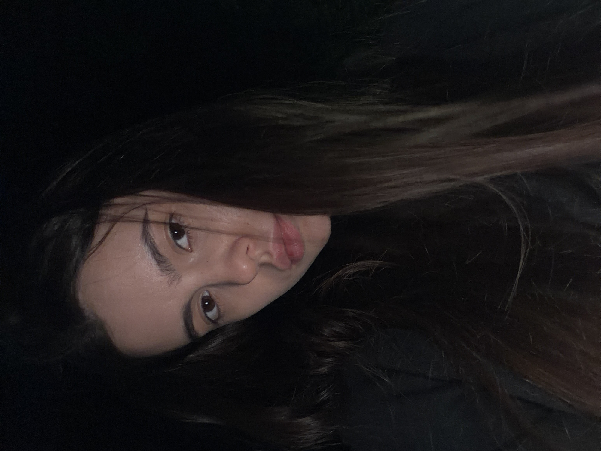 A close-up, dimly lit portrait shows a young woman with long, dark brown hair. Her face is angled towards the viewer, and her gaze is directed at the camera. Her facial features are visible: eyes, nose, and lips. The background is dark, rendering the surroundings indistinct. There is no information given in the image or the text to indicate the location. Therefore, it's impossible to determine if the location is Chaikovsky, Russia.