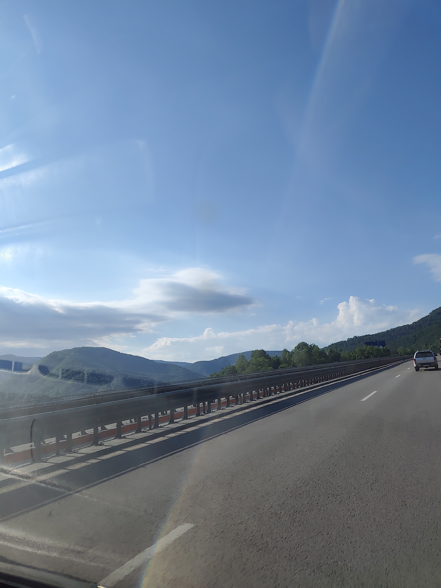 The image shows a view from a vehicle traveling on a highway. The highway has two lanes and a metal guardrail separating the lanes from the embankment. The embankment is covered with greenery and trees, with mountains in the background. The sky is blue with some clouds. A white SUV is visible on the highway ahead. The image is likely taken in Vrachesh, Bulgaria, based on the presence of a highway and mountainous terrain typical of the region.