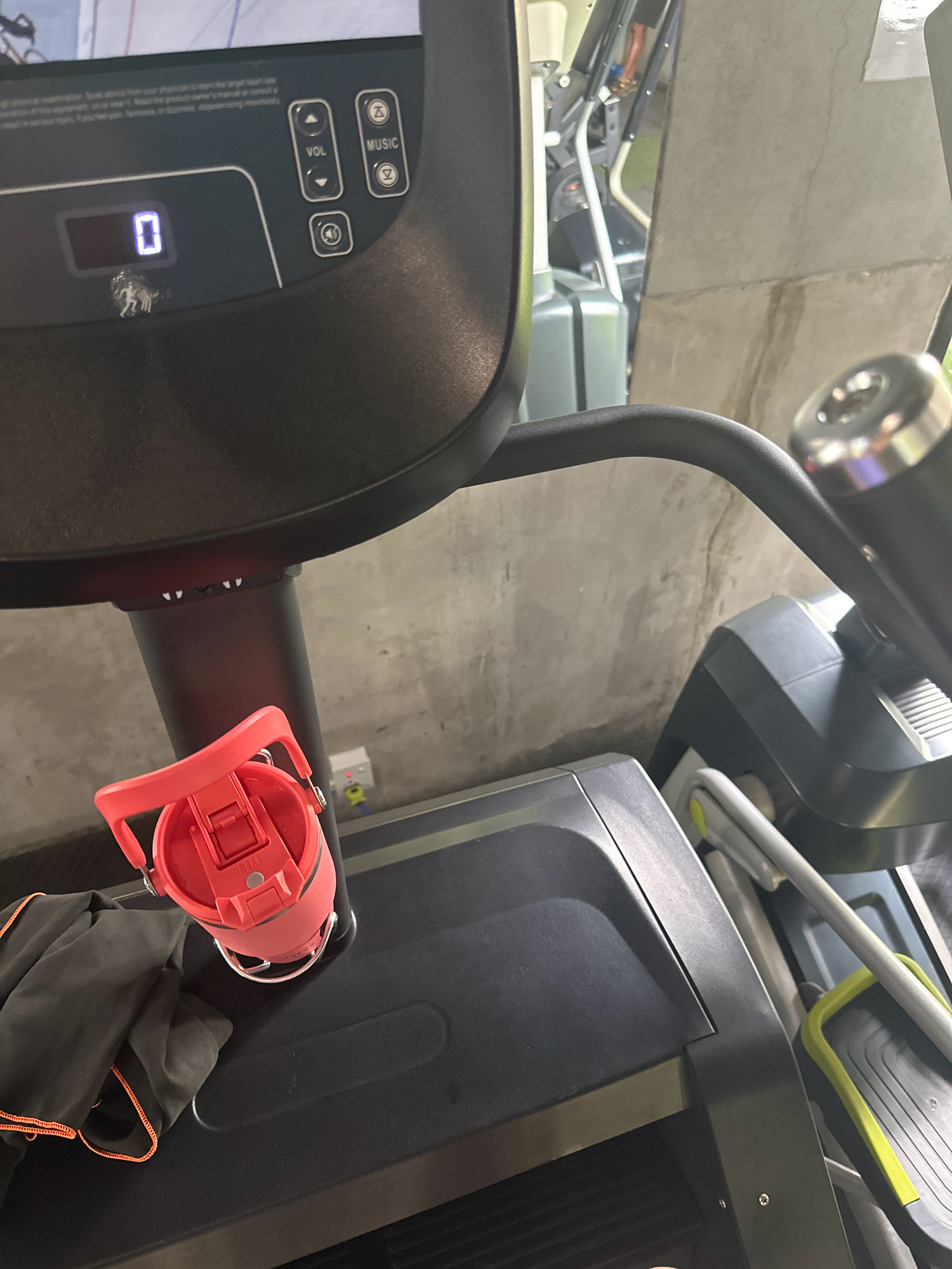 The image shows a close-up view of a portion of a treadmill console and the area around it. The console features a digital display showing the number "0", along with buttons for volume and music control, and a speaker icon. Below the display, a small metallic figurine of a horse is attached. To the left of the console, a bright red water bottle with a white lid and a handle is placed on the treadmill's deck. A dark grey towel with orange stitching is also visible on the deck, partially underneath the water bottle. In the background, a portion of another gym machine with grey and green elements can be seen, and the wall behind appears to be made of concrete. The lighting suggests an indoor environment, likely a gym. The scene is set in a fitness facility in Lalitpur, Nepal.
