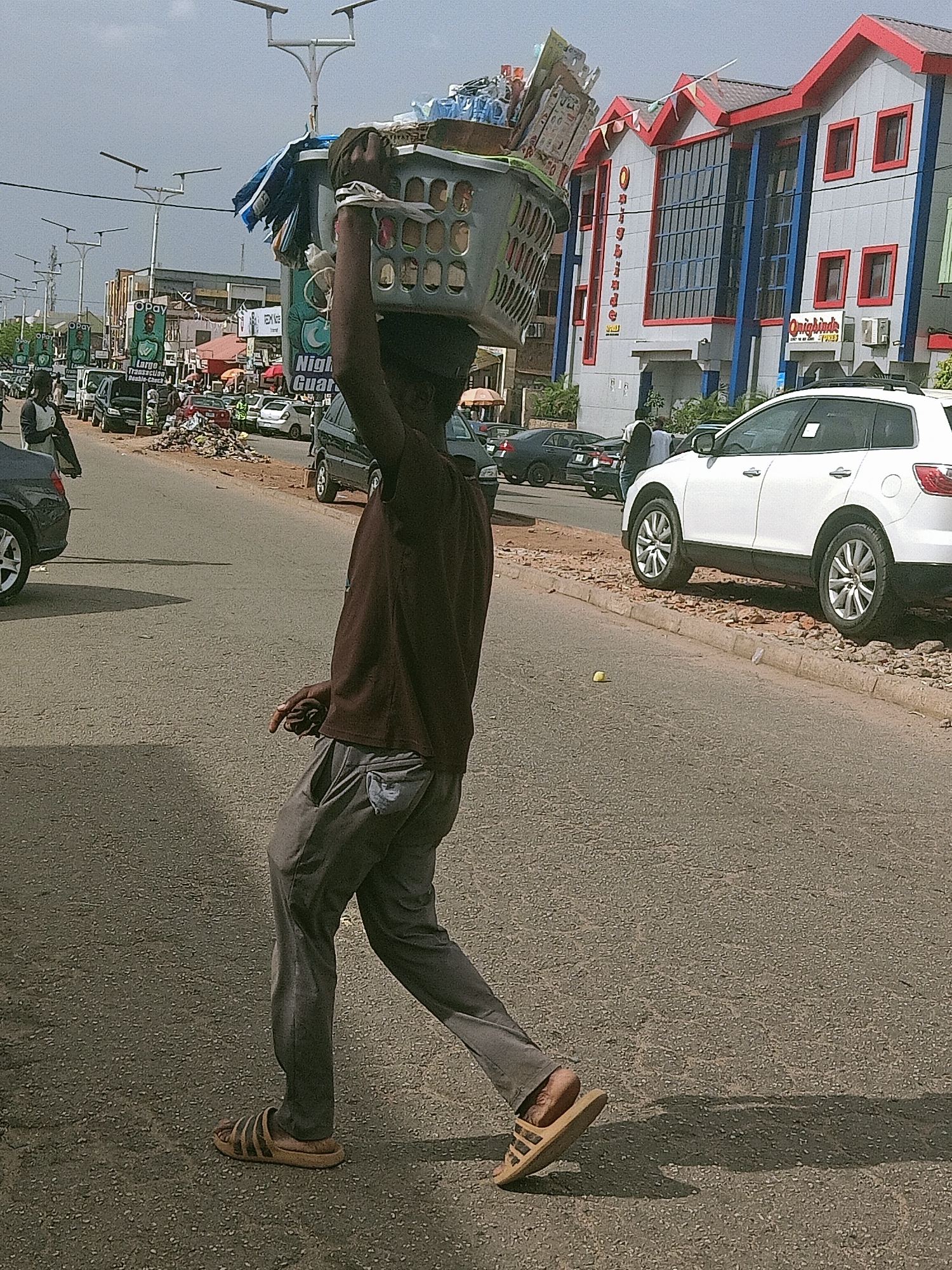 A street scene in Jos, Nigeria, depicts an individual walking on the road carrying a basket on their head filled with various items. The person is wearing a brown shirt, gray pants, and sandals. In the background, there are cars, buildings, and streetlights. The "Onigbinde's" store is visible. A white SUV is parked on the side of the road.
