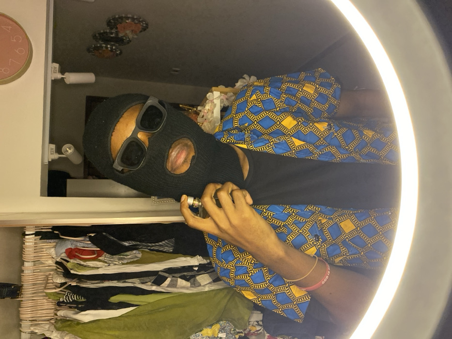 An individual is depicted indoors, likely within a private residence in Tema, Ghana, positioned in front of a ring light. The person, whose gender is not definitively ascertainable, wears a black balaclava covering the head and face, with rectangular black sunglasses obscuring the eyes. The mouth opening reveals visible lips. The individual is dressed in a black t-shirt beneath an unbuttoned, short-sleeved shirt featuring a blue and yellow geometric patterned fabric. The left wrist displays two thin bracelets, one light-colored and another reddish-pink with faint white text. The right hand holds a small, clear glass spray bottle with a metallic top, held near the left side of the balaclava-covered face as if applying its contents. The background on the left shows an open wardrobe or closet filled with various hanging garments. To the right, a dark wall is visible, adorned with a partial pink circular clock and three circular, patterned wall decorations. Overhead, white track lighting fixtures are mounted. The bright illumination from the ring light frames the lower portion of the individual, indicating the image was captured directly facing the light source.